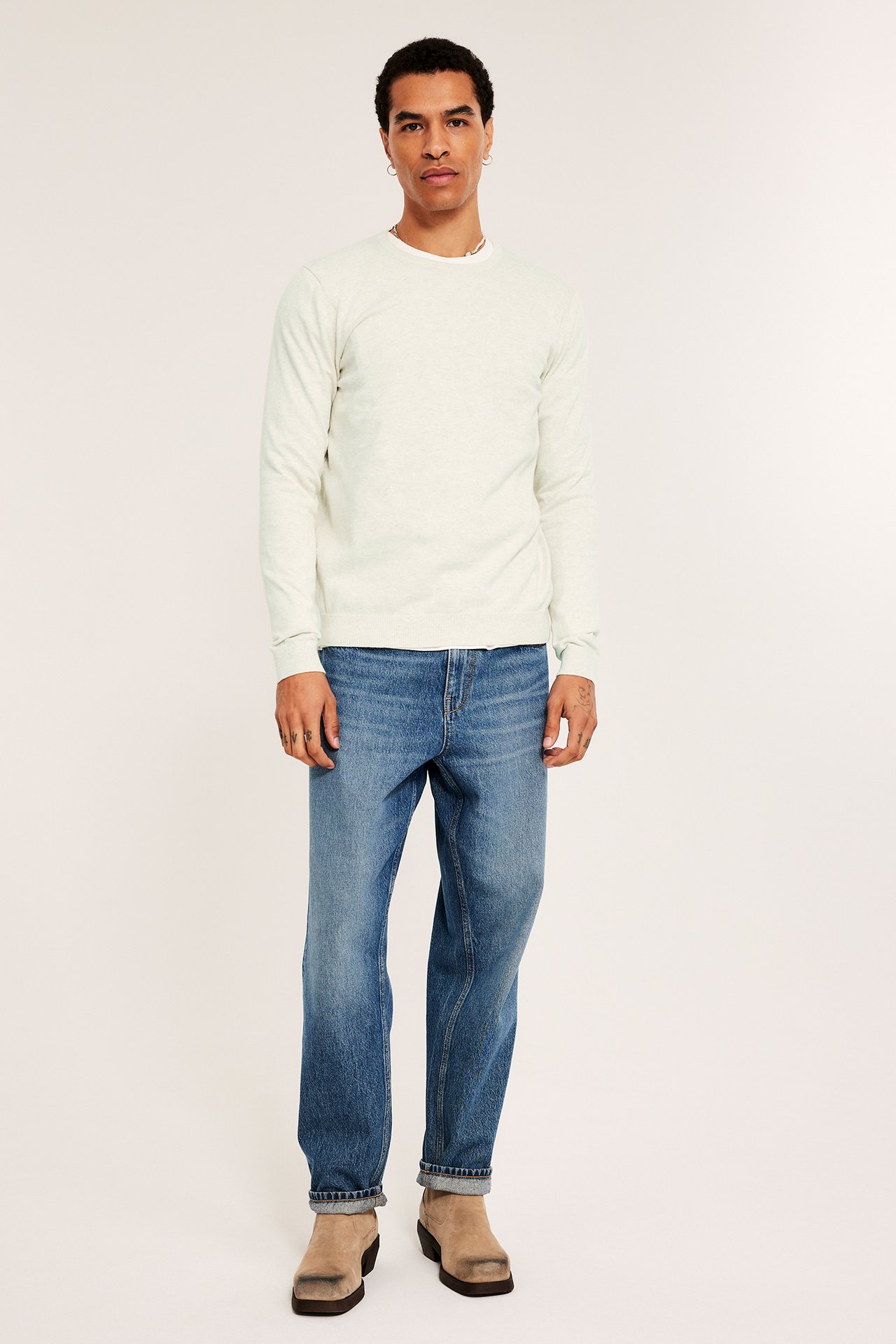 Edestä kuvattu mies, yllään valkoinen meleerattu hienoneulottu puuvillapusero, siniset farkut, beiget kengät.