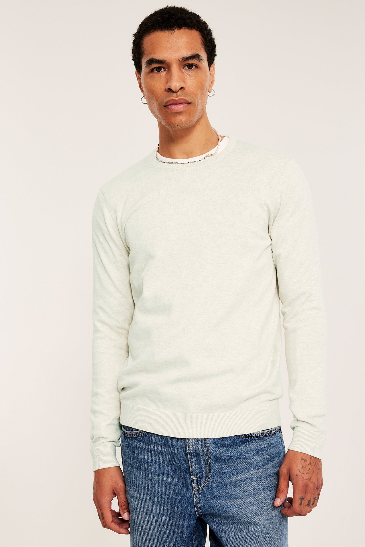 Edestäpäin: mies yllään vaalea meleerattu puuvillaneulepaita valkoisen t-paidan ja sinisten farkkujen päällä.