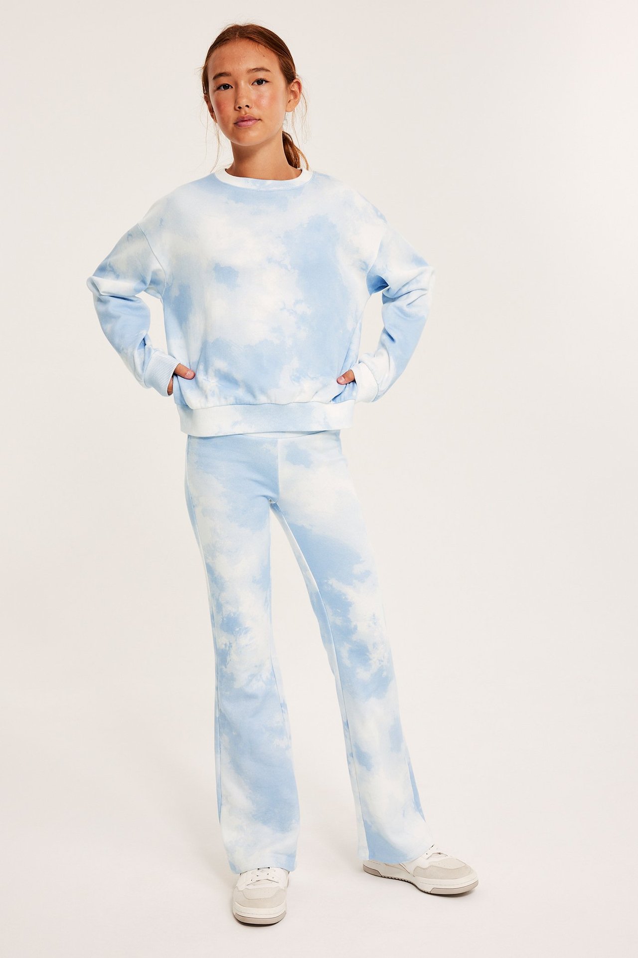 Lapsi edestä: luonnonvalkoinen ja vaaleansininen tie-dye collegepaita, levenevät housut ja lenkkarit.