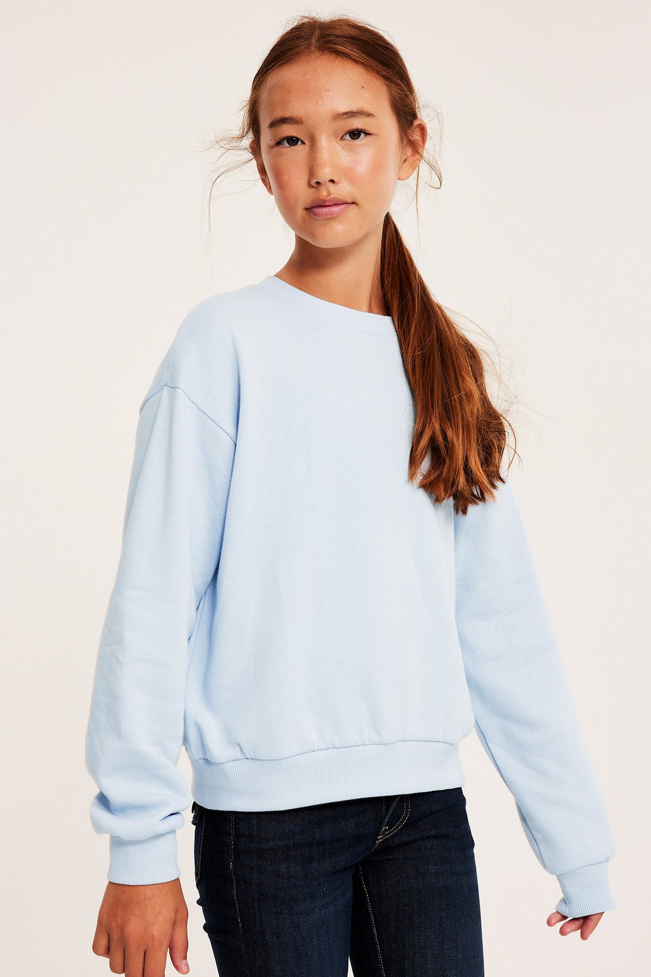 Framifrån, ett barn i en ljusblå sweatshirt med rund hals, ribbade muddar och mörka jeans.