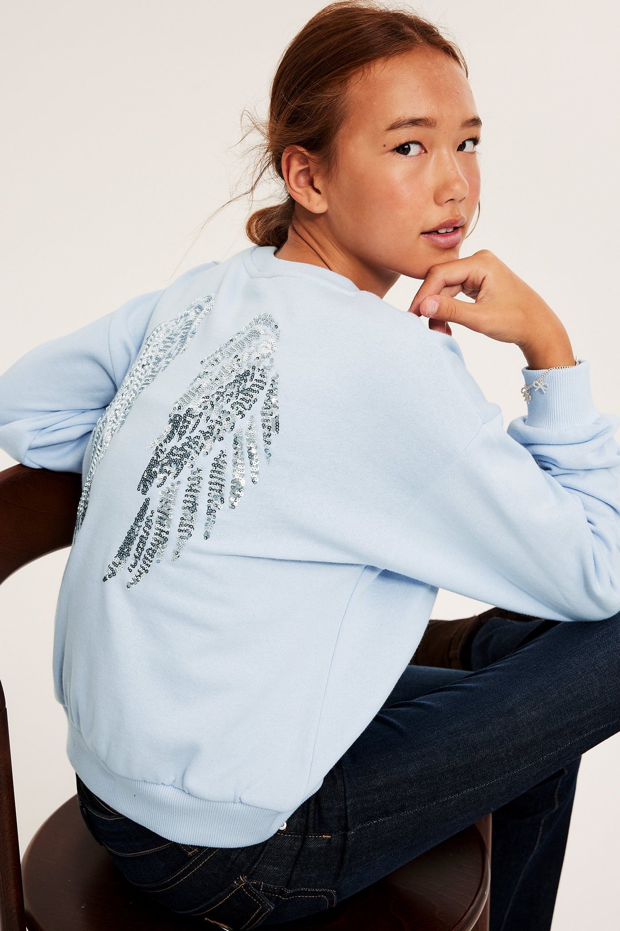 Widok z tyłu: dziecko w jasnoniebieskiej bluzie z cekinowymi skrzydłami i ciemnoniebieskich dżinsach.