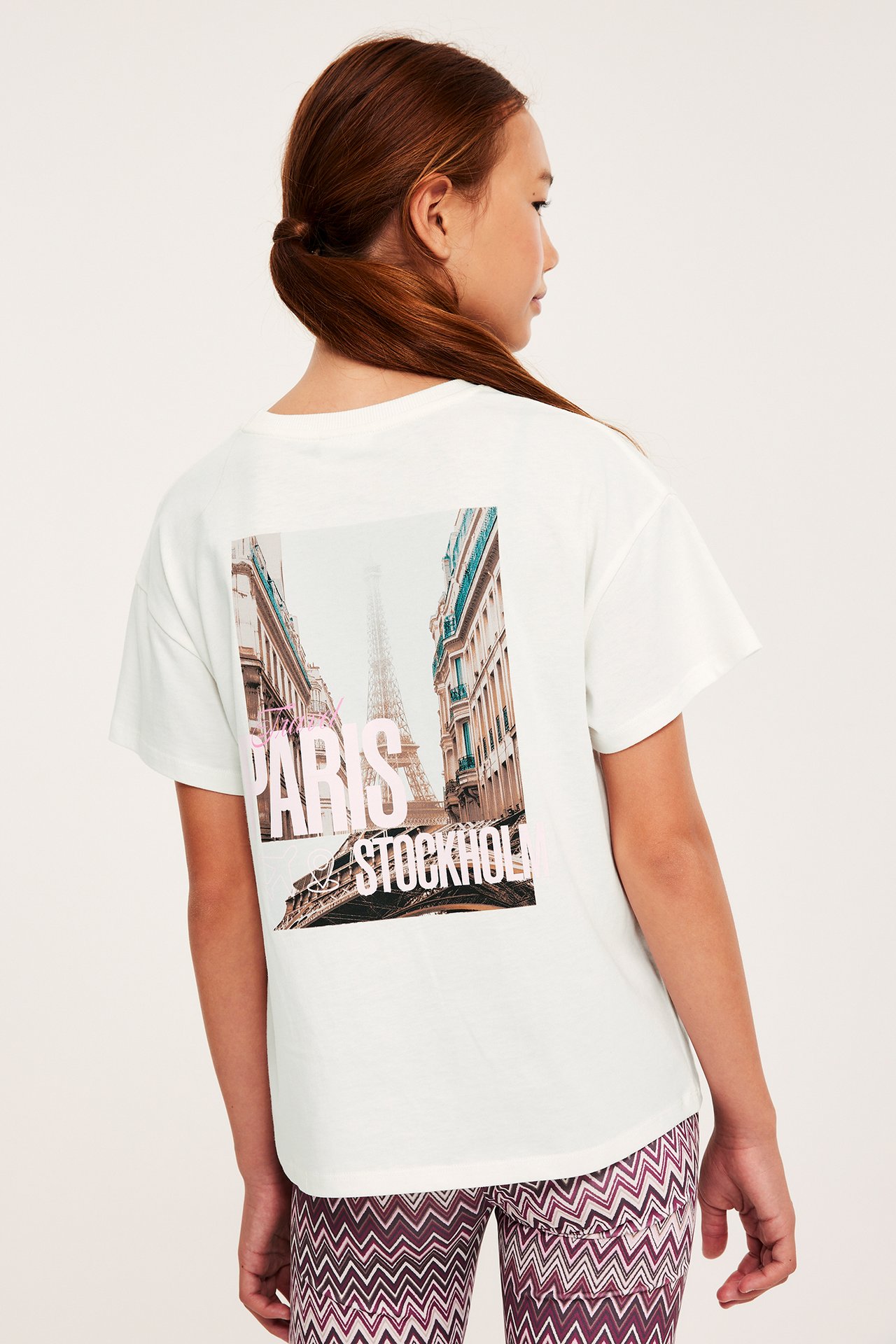 Bakfra av et barn i en offwhite bomulls-T-skjorte med Paris/Stockholm-trykk og sikksakkmønstrede leggings.