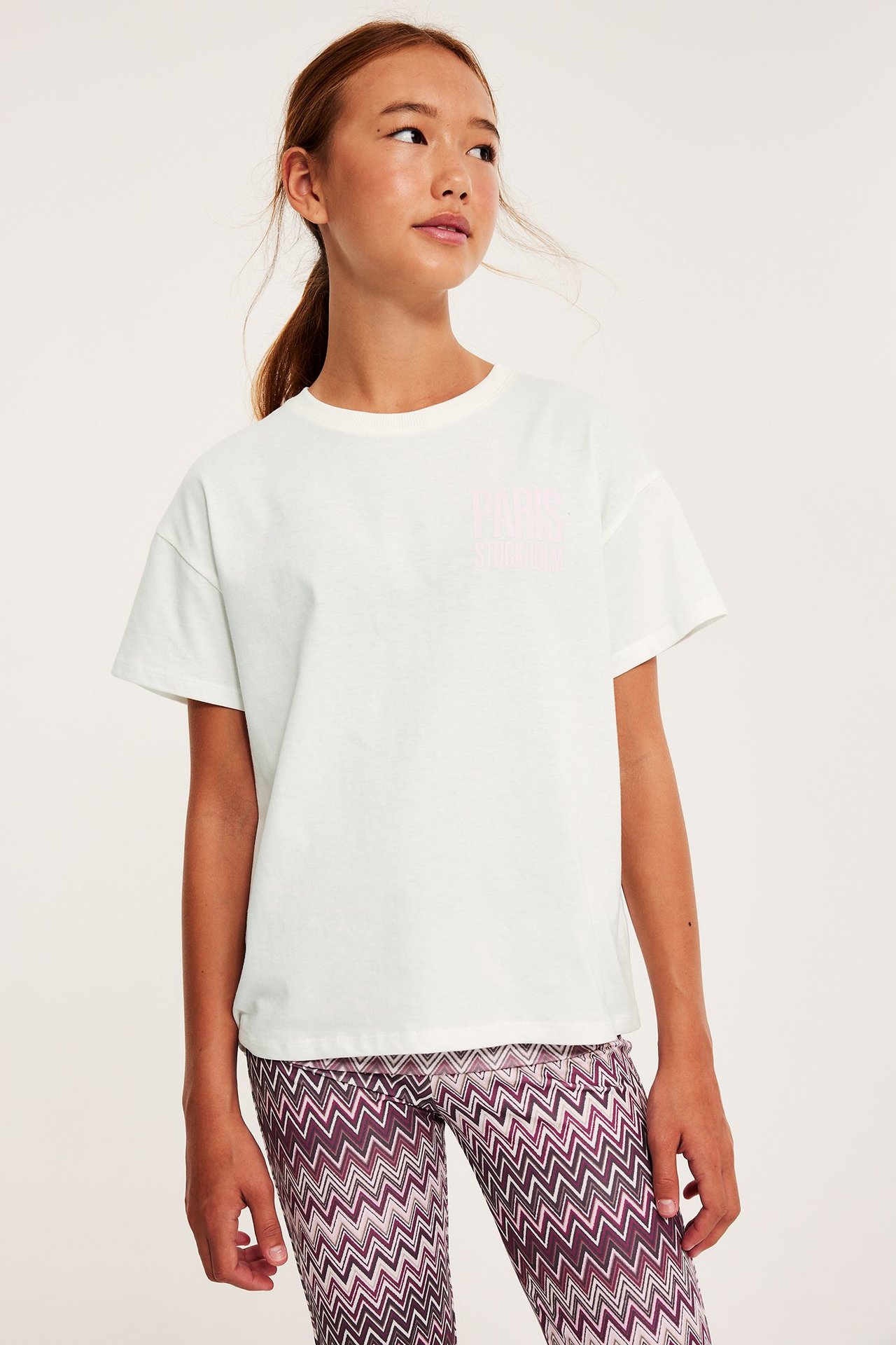 Frontvy: Flickbarn i offwhite bomulls-t-shirt med rosa tryck fram och mönstrade leggings.