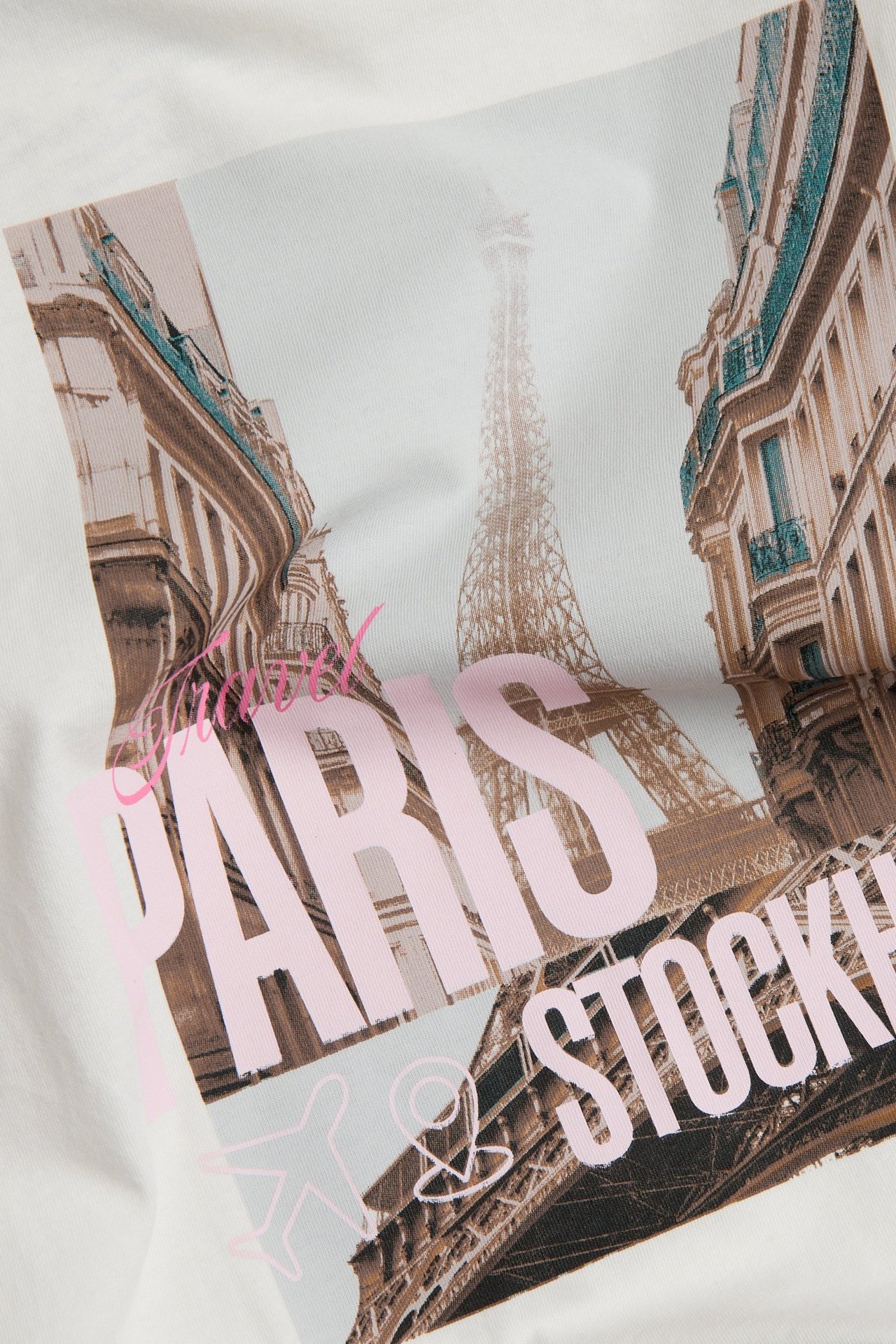 Zbliżenie: dziecięca koszulka bawełniana off-white z wieżą Eiffla, paryskimi budynkami i różowym napisem 'PARIS & STOCKHOLM'.