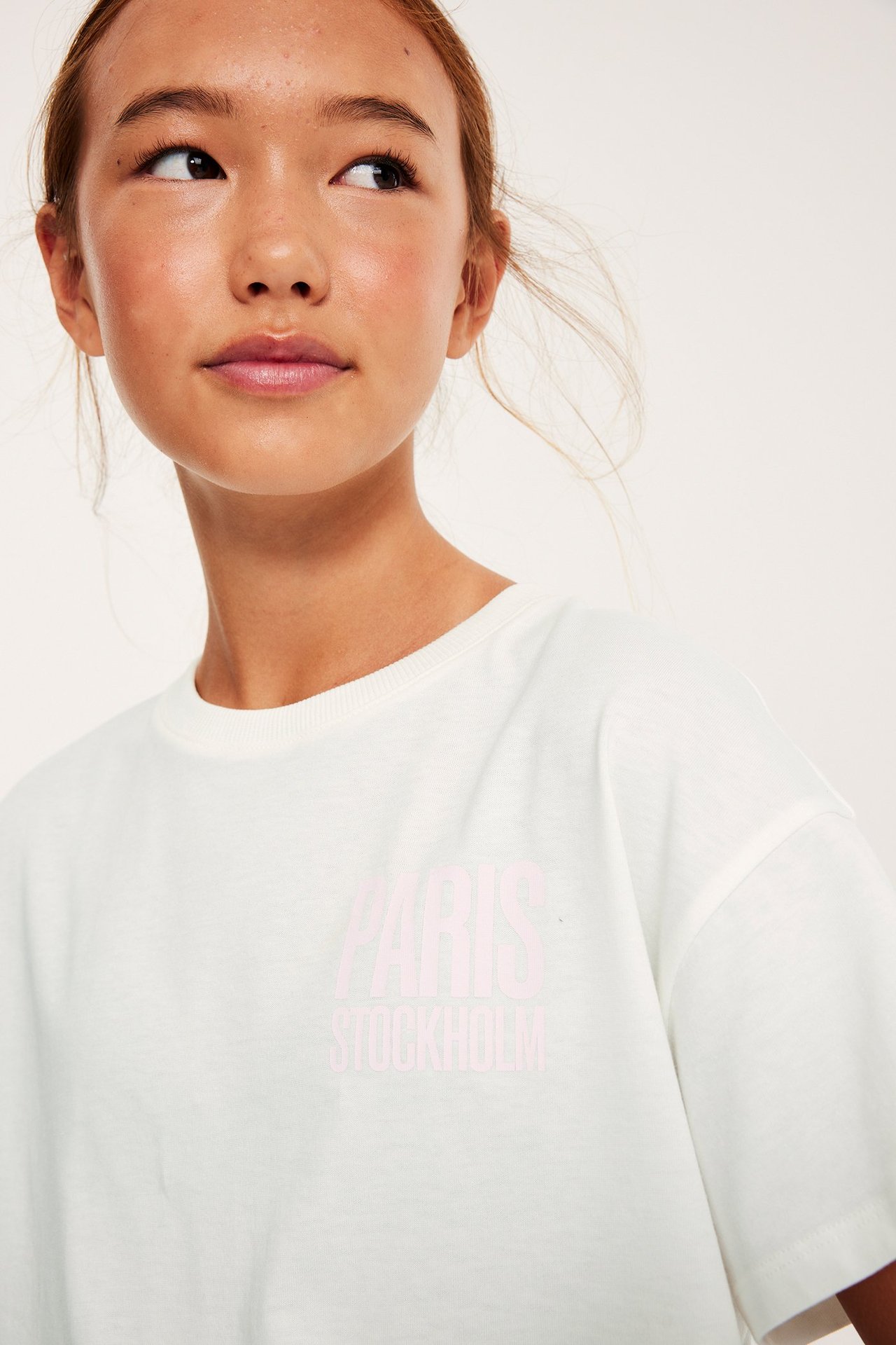 Närbild framifrån på ett barn i en offwhite bomulls-t-shirt med ljusrosa 'PARIS STOCKHOLM' tryck.