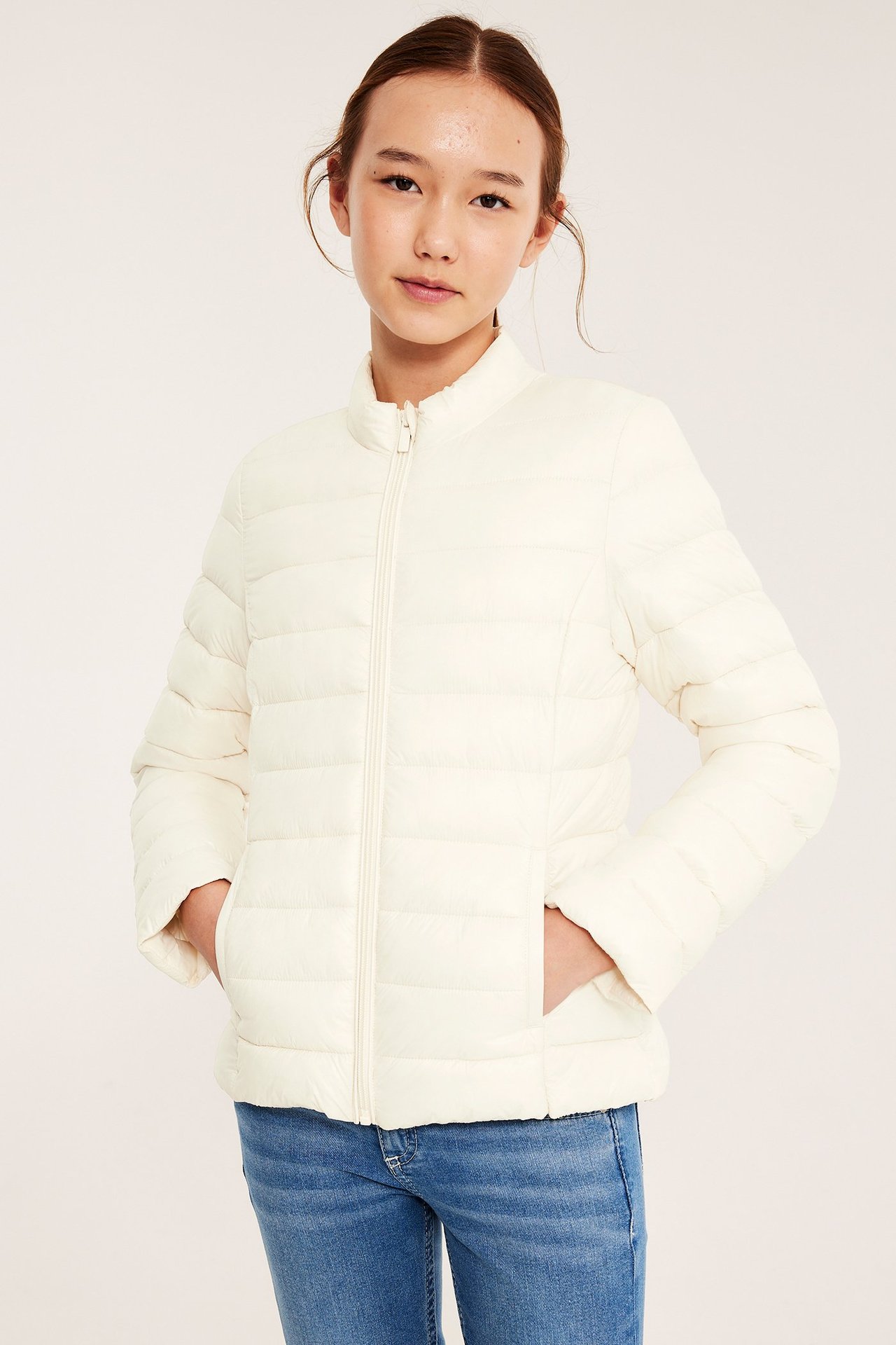 Edestä: Lapsi pukeutunut luonnonvalkoiseen kiiltävään toppatakkiin vetoketjulla, kädet taskuissa, ja siniset farkut.