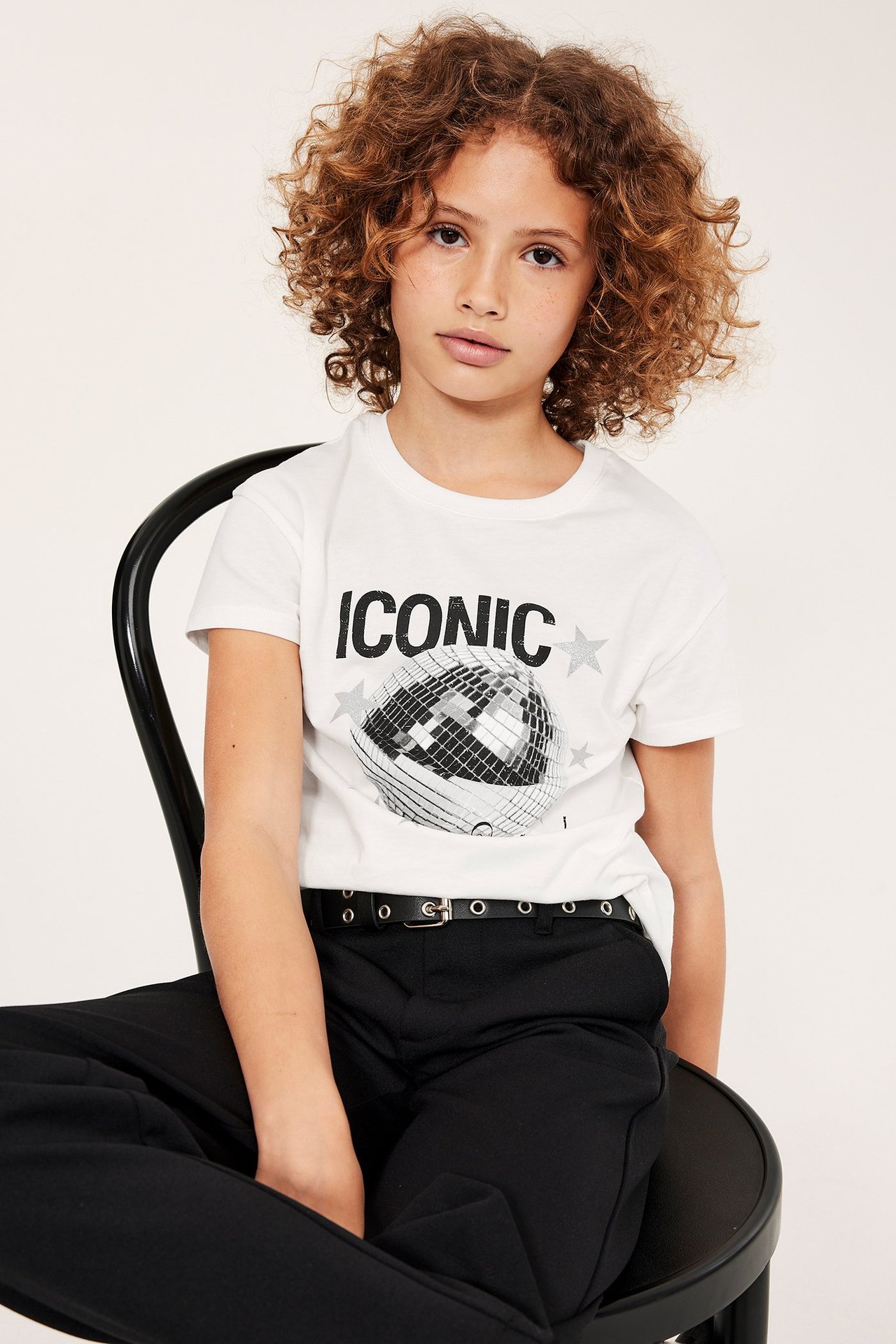 Framifrån: Barn i offwhite T-shirt med 'ICONIC' discokulstryck, svarta byxor och svart bälte.