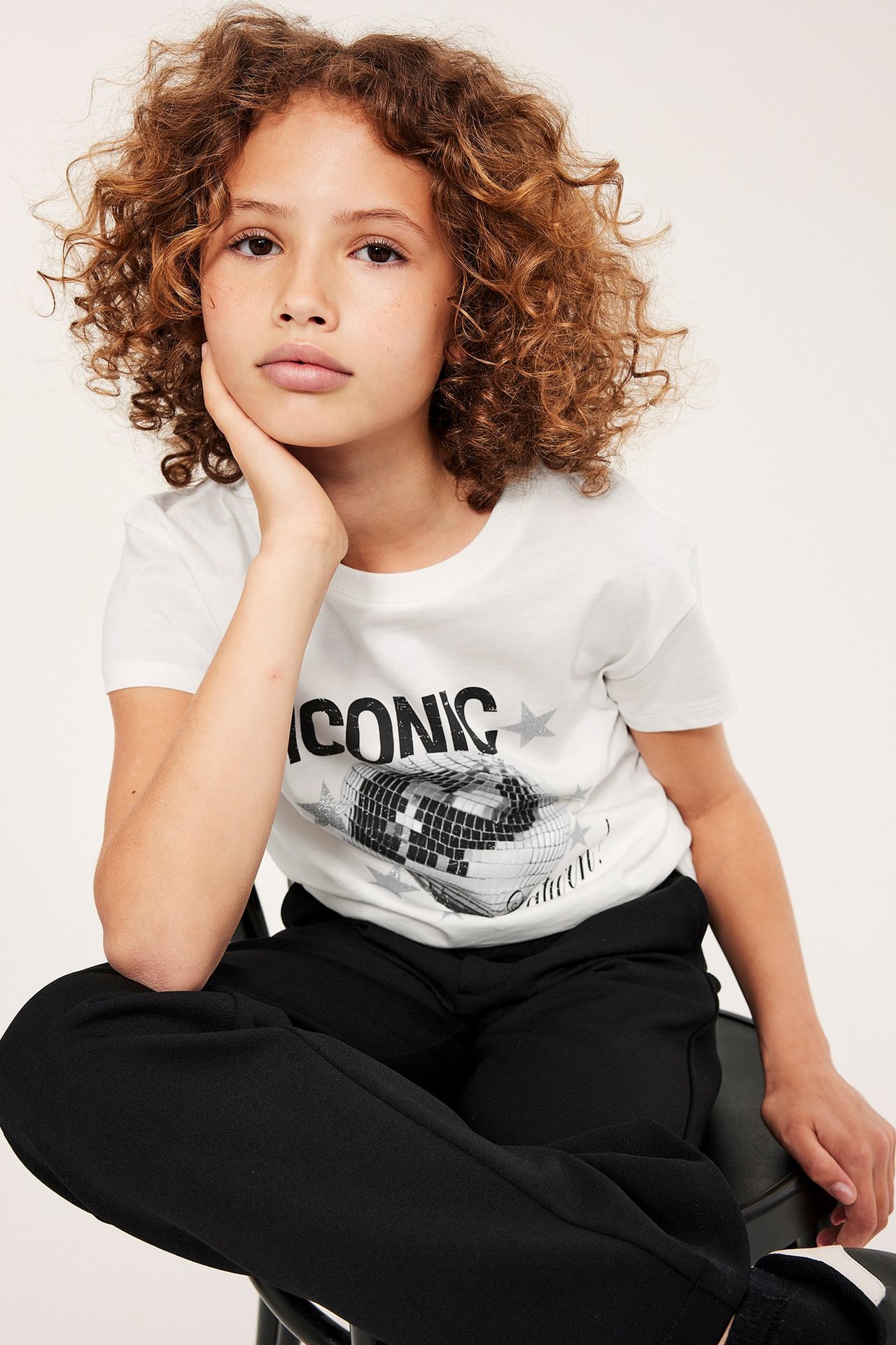 Frontvy av ett barn i en offwhite bomulls-T-shirt med "ICONIC" discobollstryck och svarta byxor.