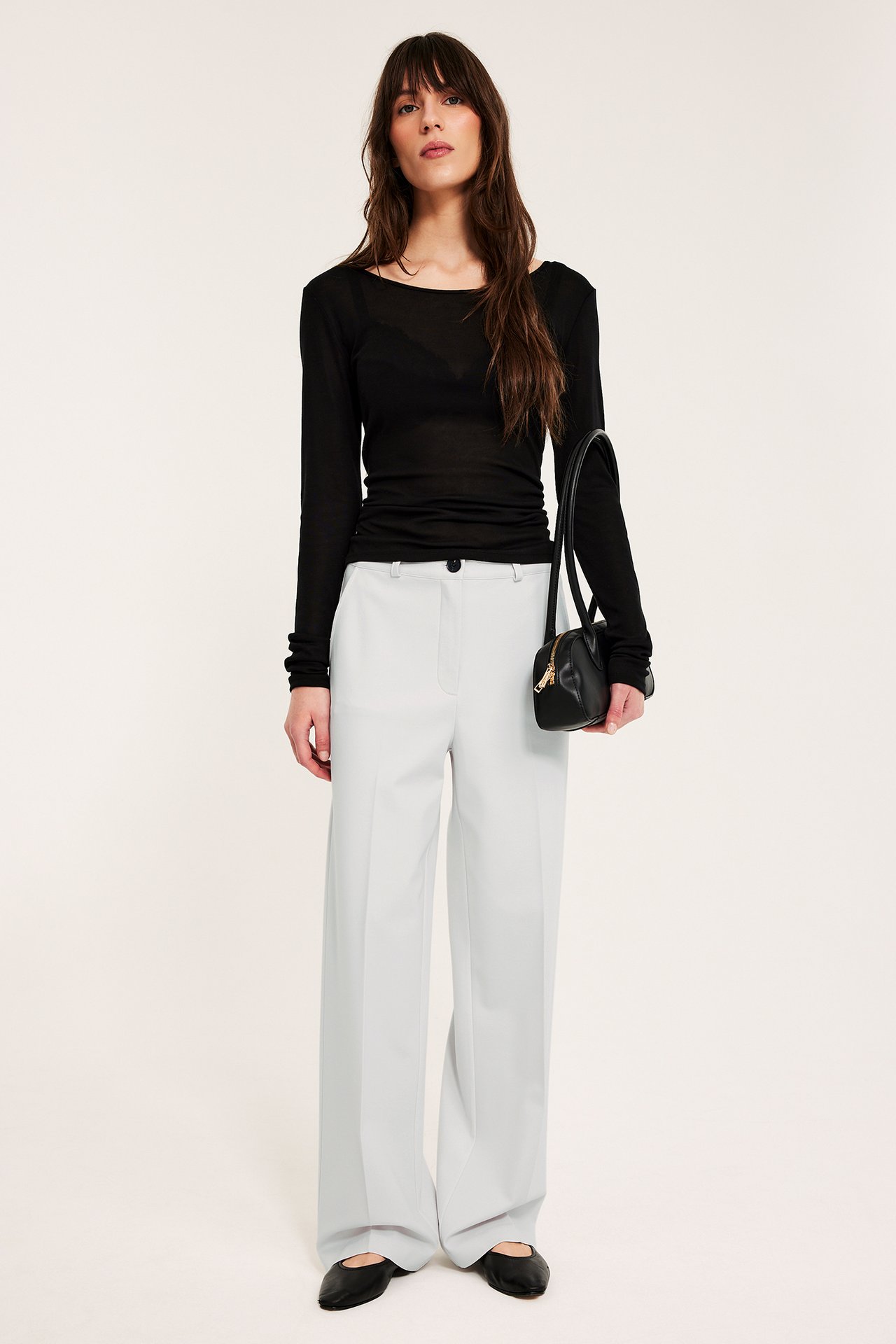 Frontbilde av kvinne i hvite vide dressbukser, svart gjennomsiktig langermet topp, holder svart håndveske.