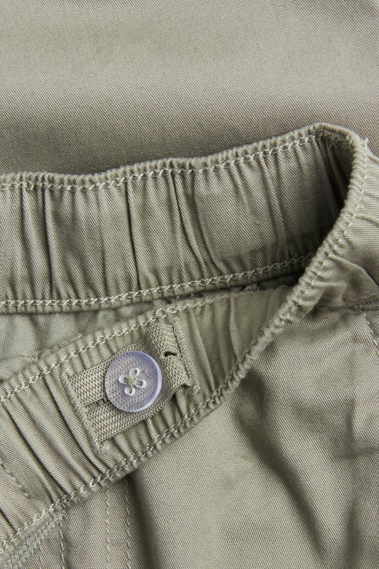 Lähikuva khakinvihreistä puuvillahousuista, joissa joustava vyötärö ja säädettävä nappi.