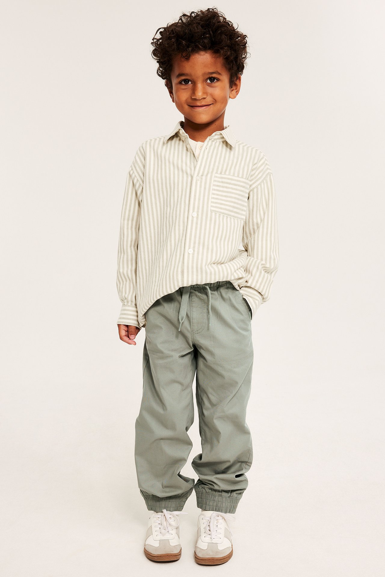 Framifrån, ett barn i khakigröna mjukisbyxor med resårmuddar, en randig skjorta och vita sneakers.