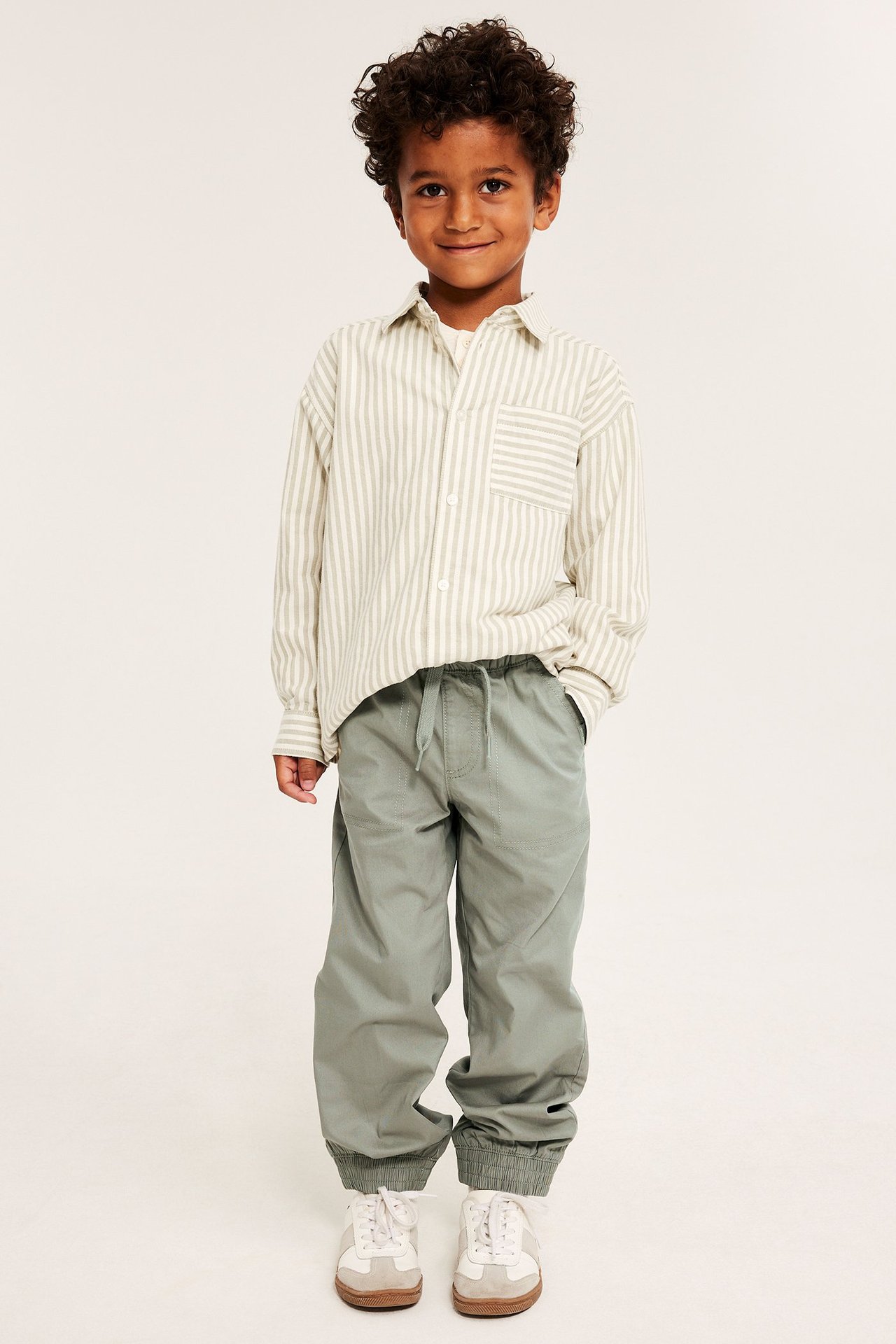 Edestä, lapsi khakinvihreissä resorilahkeisissa collegehousuissa, raidallisessa paidassa, valkoiset lenkkarit.