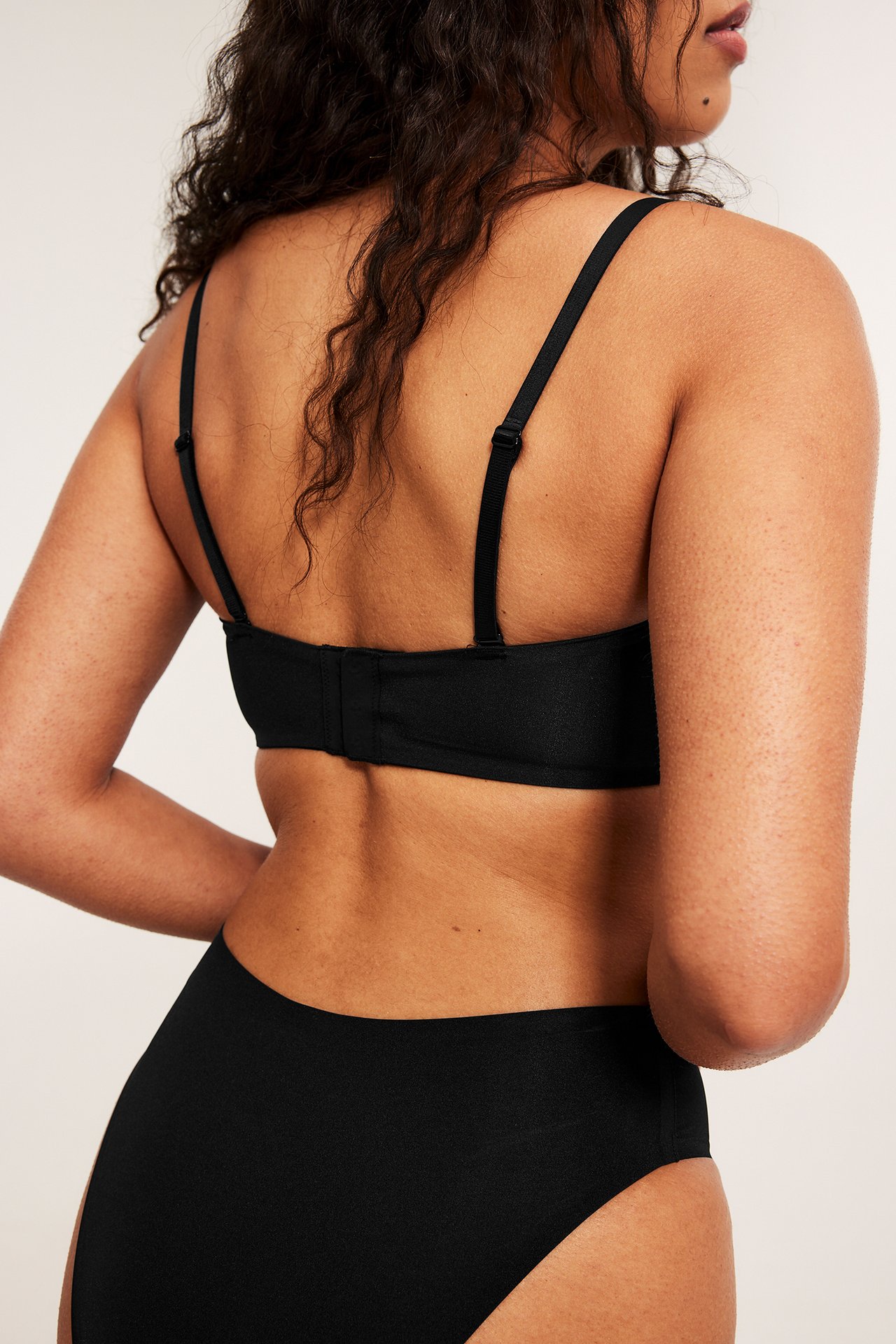 Ryggvisning av kvinne i svart bandeau-BH med justerbare stropper, hektelukking, matchende truse.