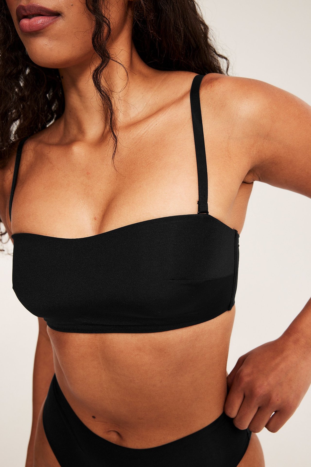 Frontvisning av en kvinne i svart polstret bandeau-bh med tynne justerbare stropper og sorte truser.
