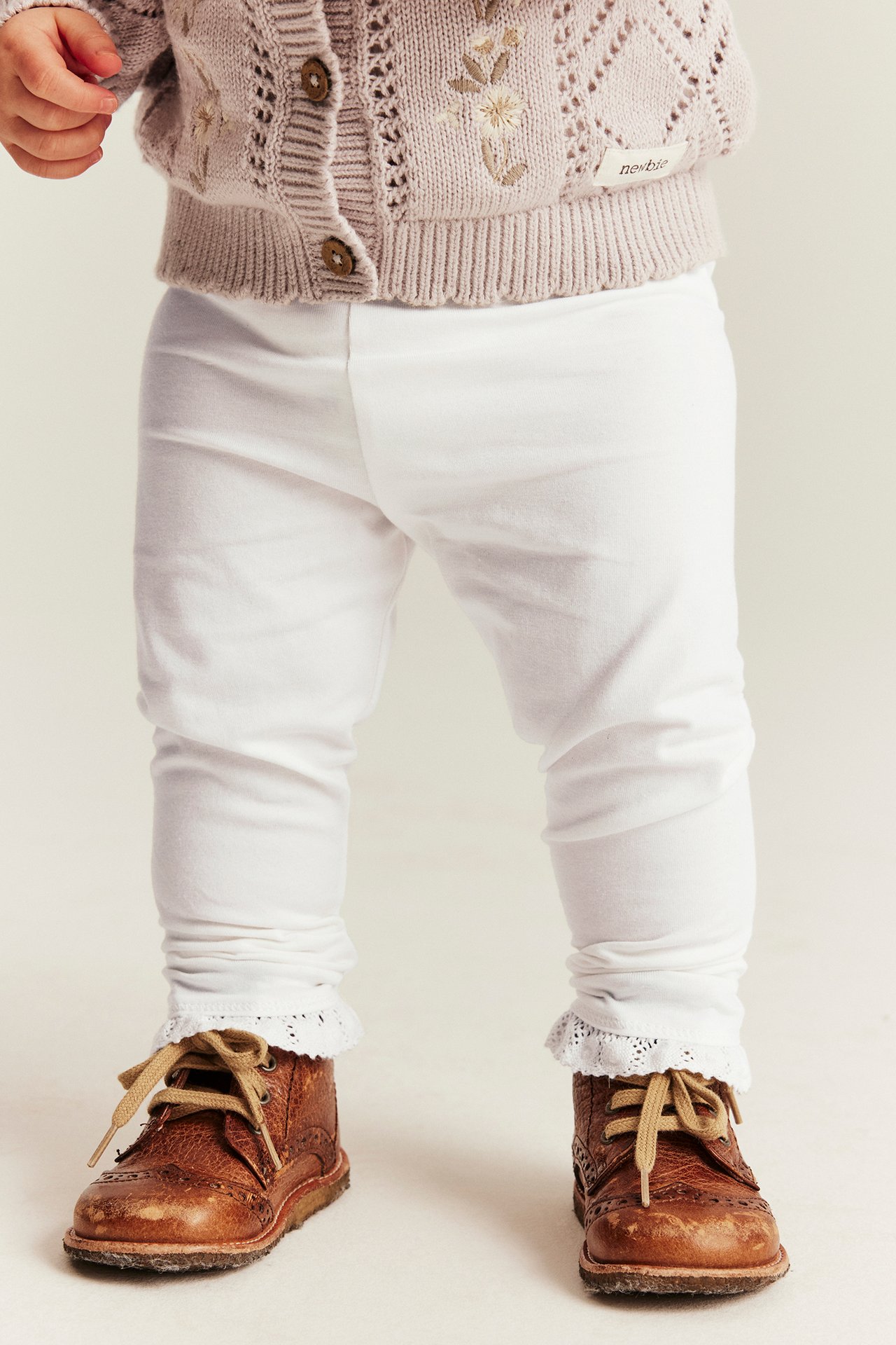 Framsida av baby i offwhite stickade leggings med spets, ljusbrun kofta och bruna skor.