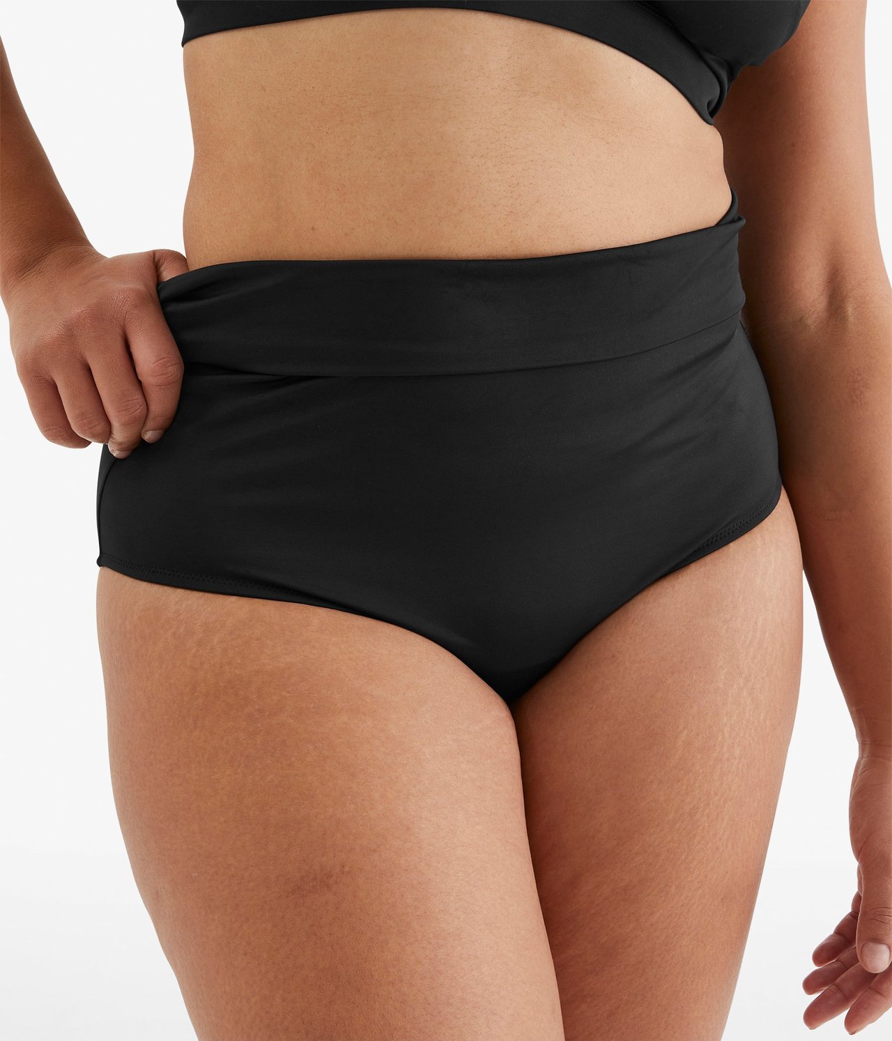 Widok z przodu: czarne, wysokie figi bikini ze składanym pasem, noszone przez kobietę. Jej ręka trzyma pas.