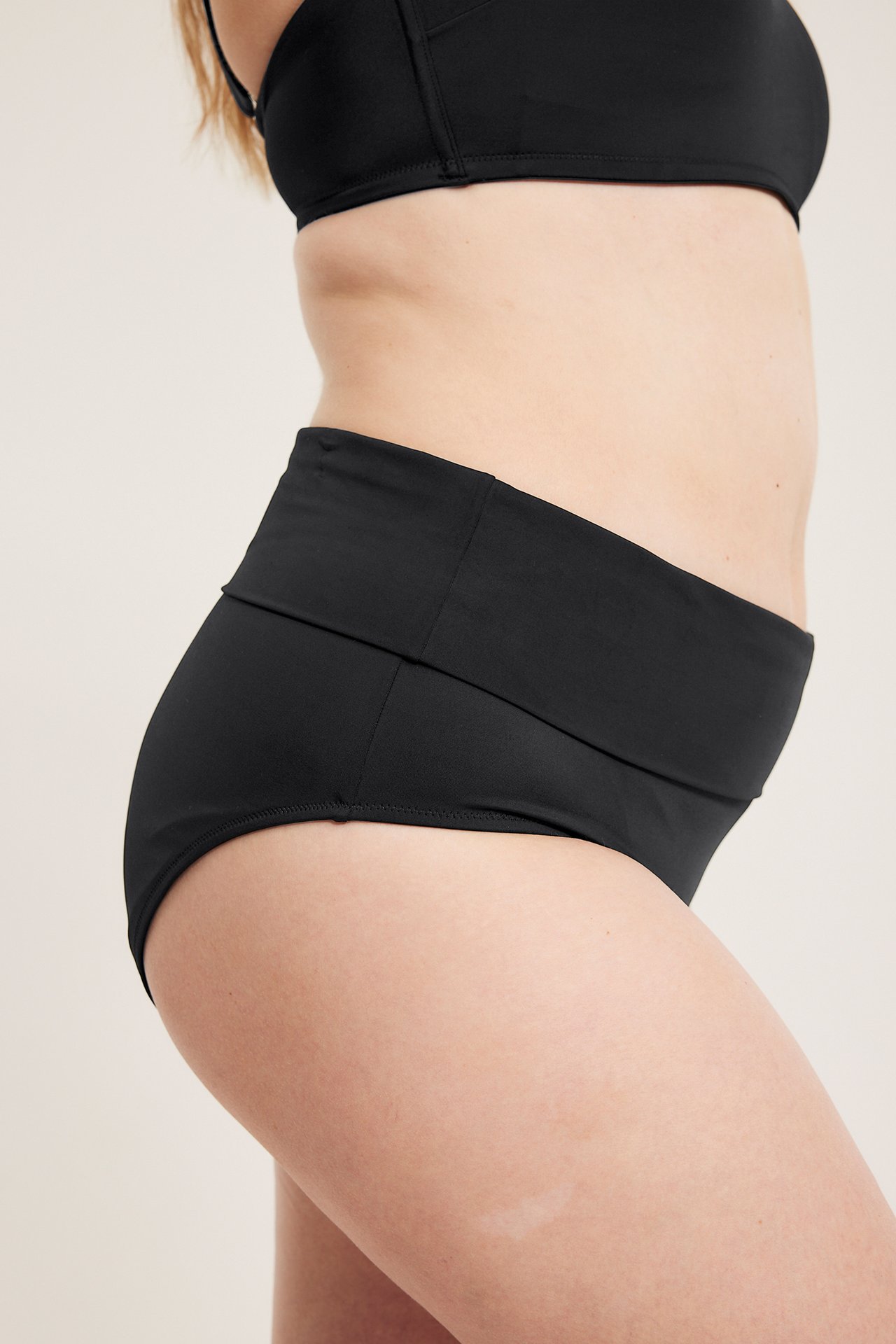 Sidevisning: Kvinne bærer svarte ensfargede bikinitruser med nedbrettbar midje og en svart bikinitopp.