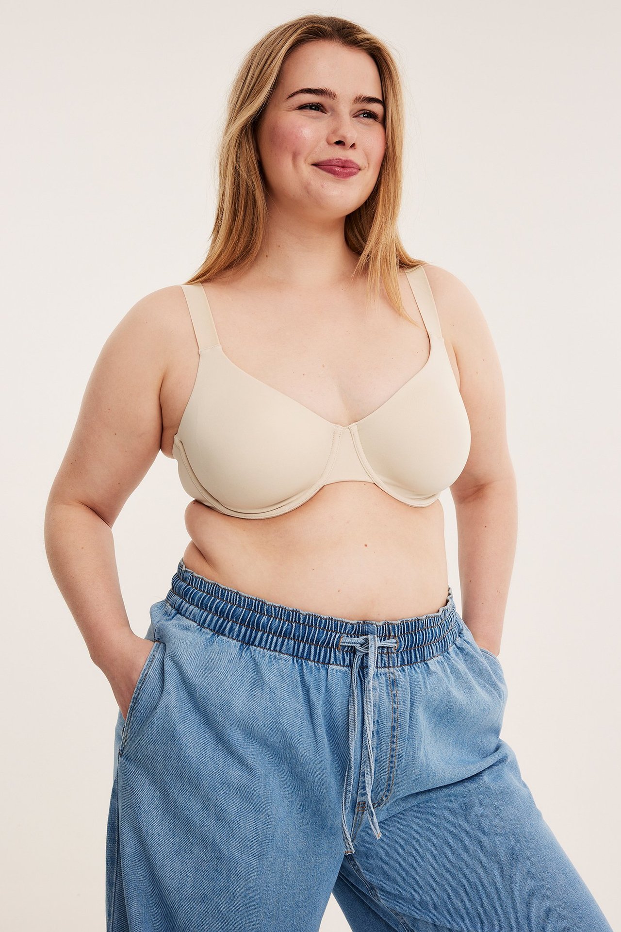 Framifrån: kvinna i ljusbeige minimizer-bh och blåa vida jeans med resår i midjan.