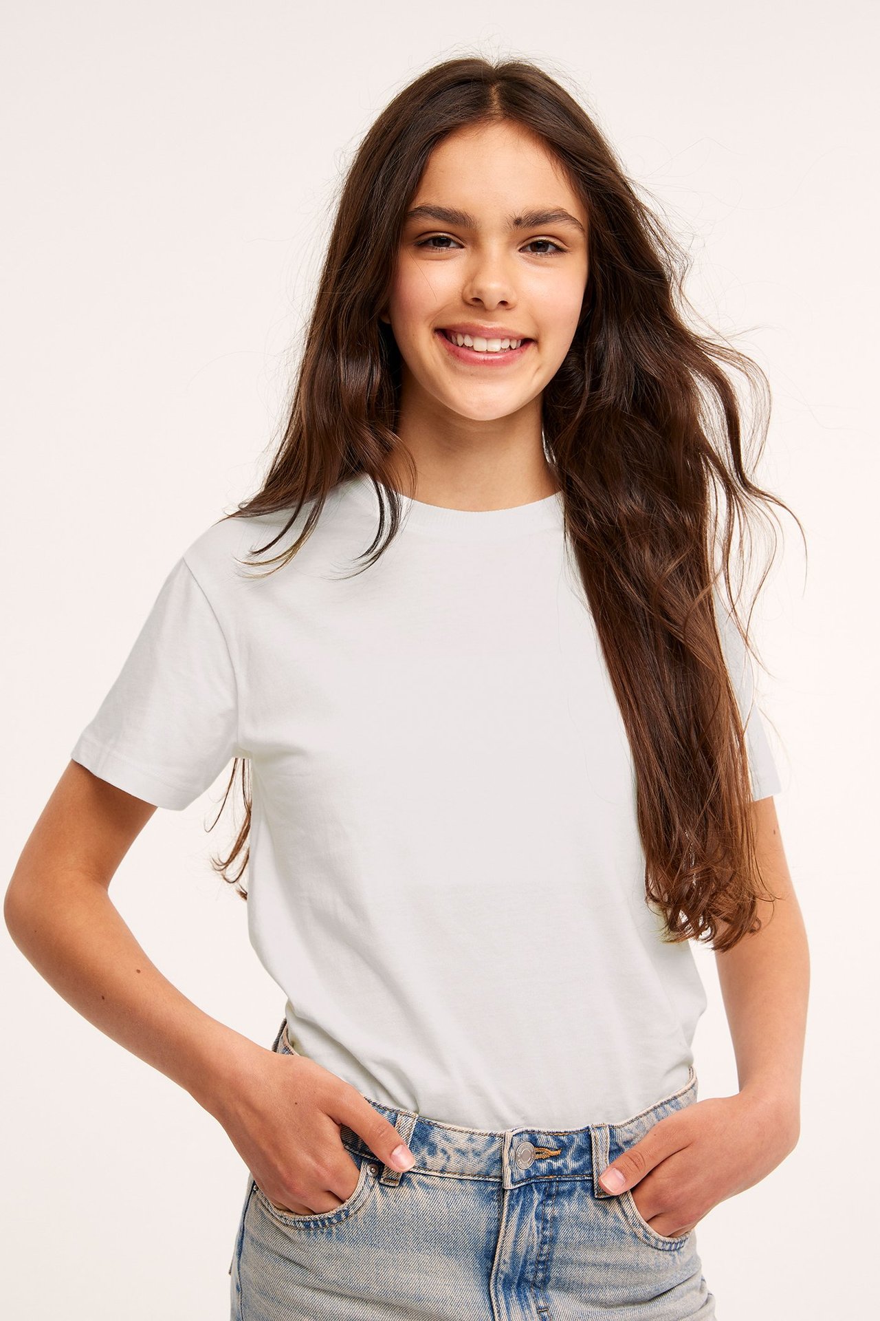 Forfra, smilende barn i offwhite bomulls-t-skjorte og lyseblå jeans med hendene i lommene.