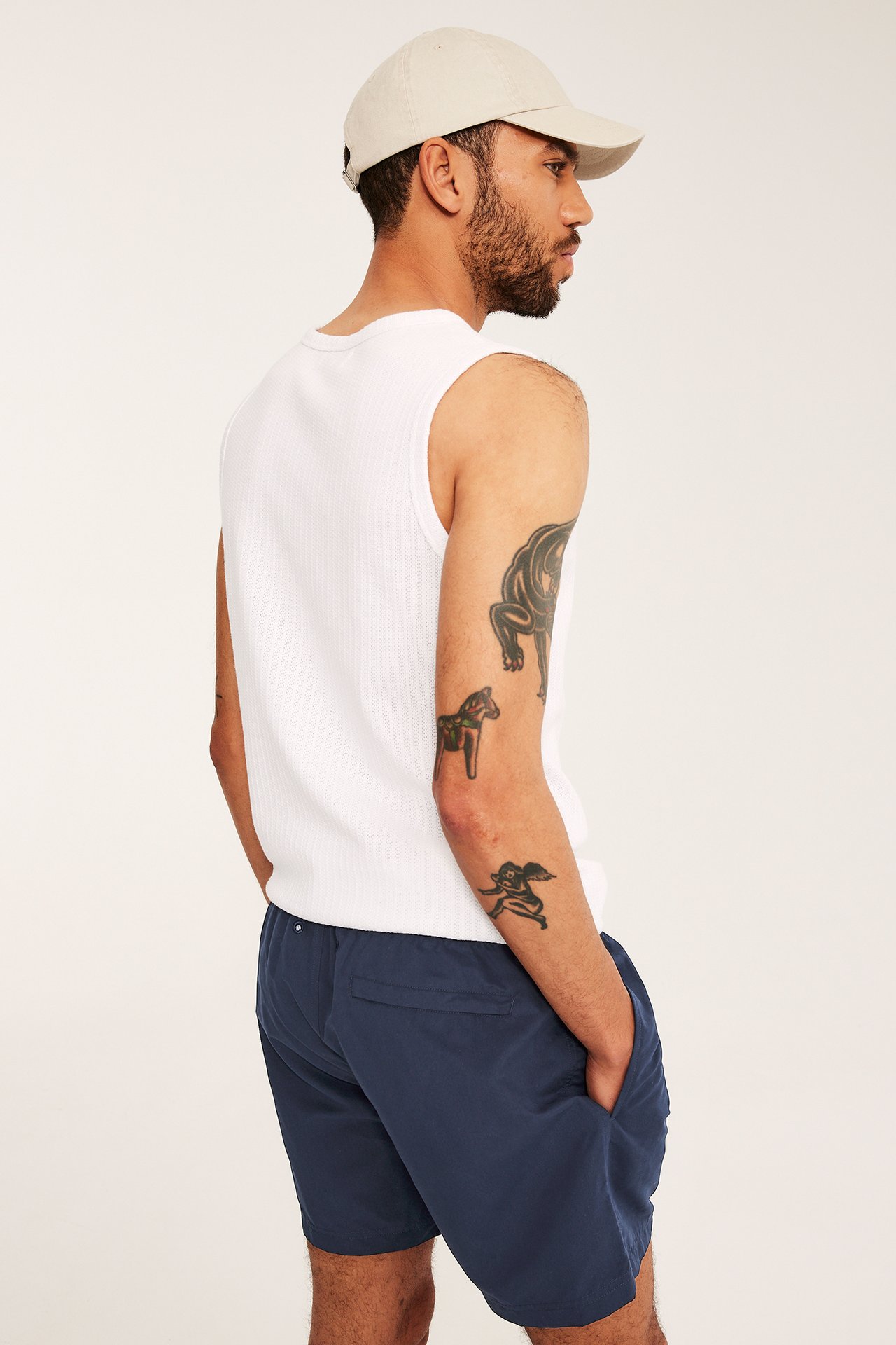Sidebilde: Mann i mørkeblå badeshorts med sidelommer, hvit ribbet topp og lys beige caps.