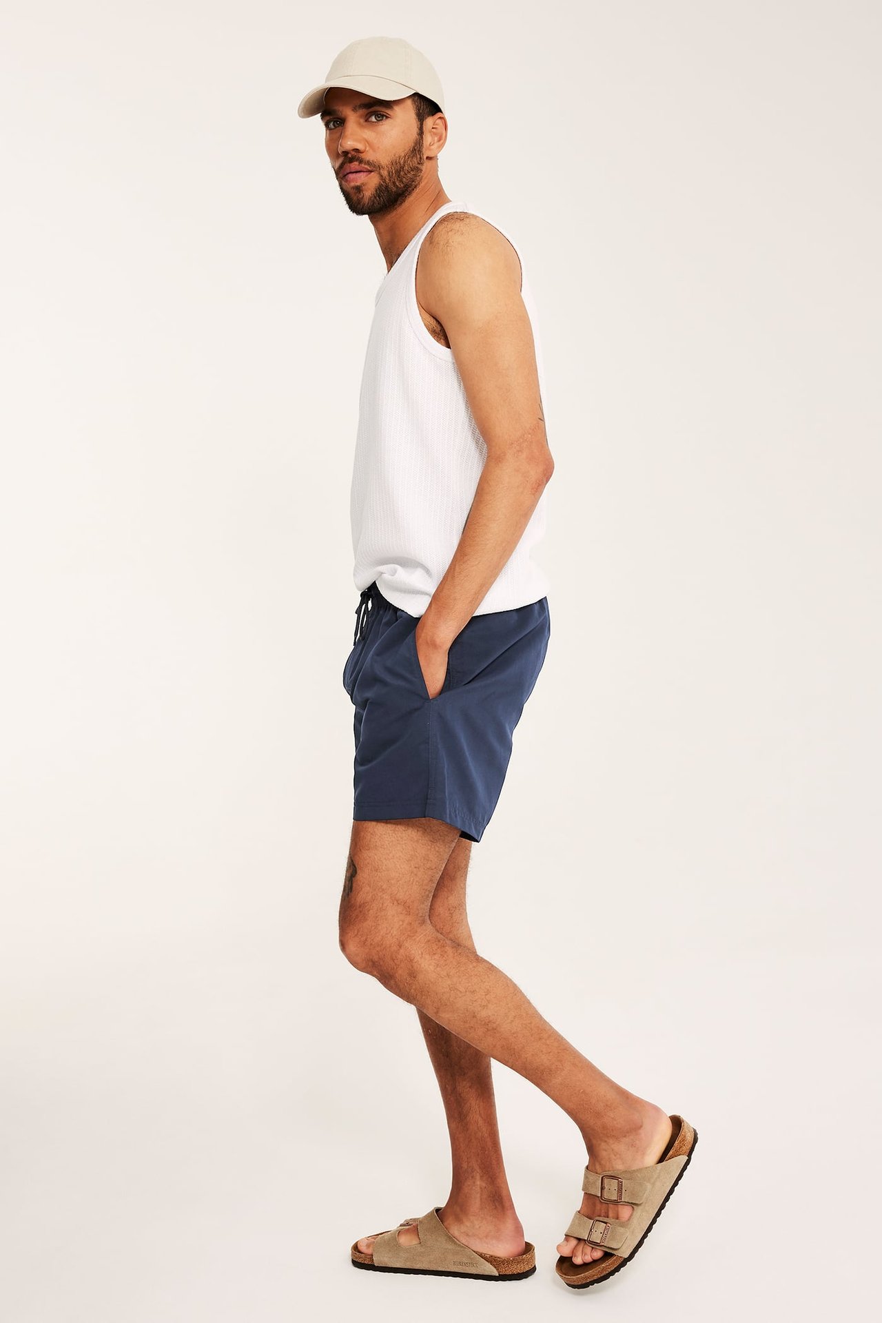 Sivukuva miehestä tummansinisissä uimashortseissa, valkoisessa ribbitoppissa, beigessä lippiksessä ja ruskeissa sandaaleissa.