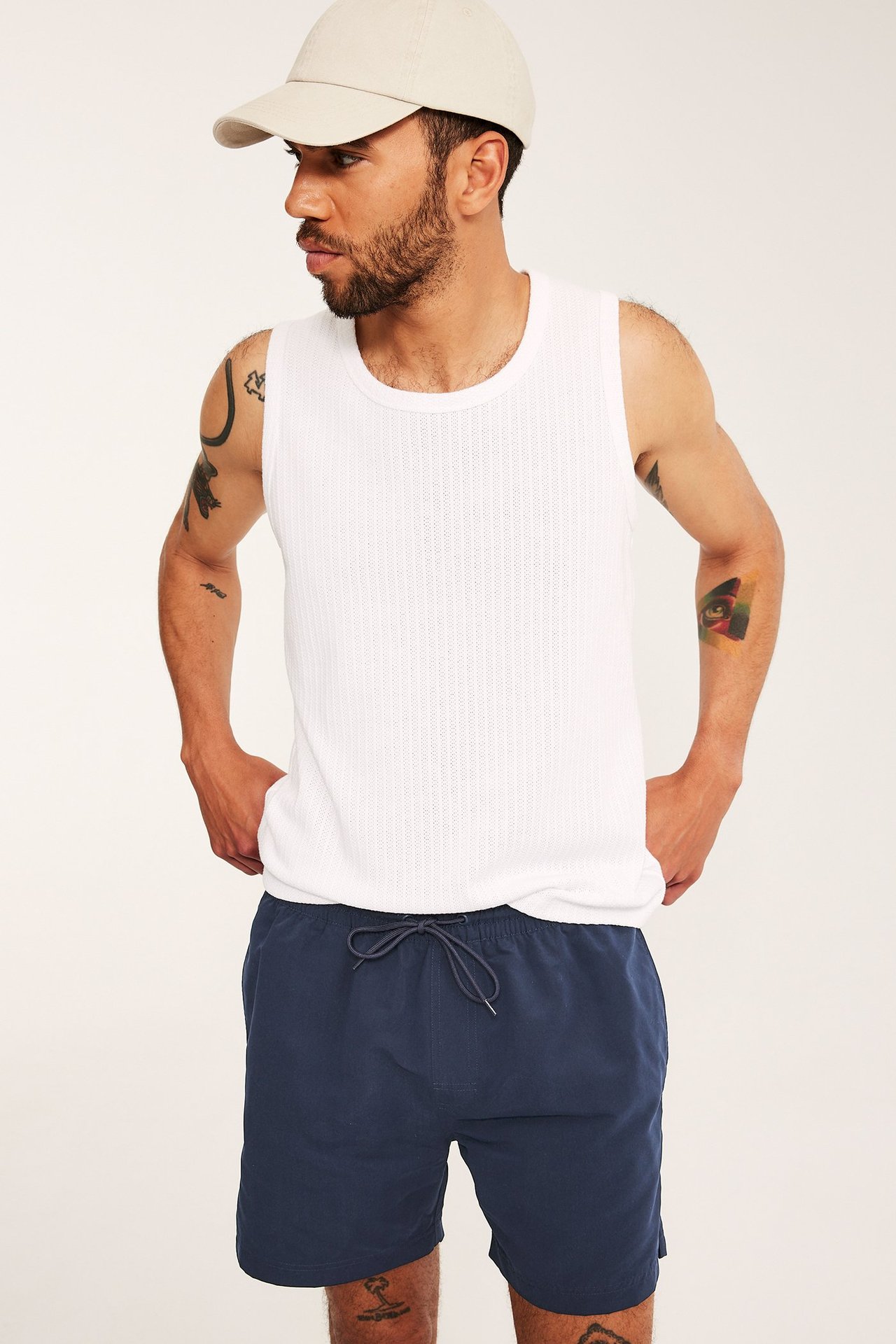 Edestä päin mies tummansinisissä uimashortseissa, valkoisessa ribbineuleisessa topissa ja beigessä lippalakissa.