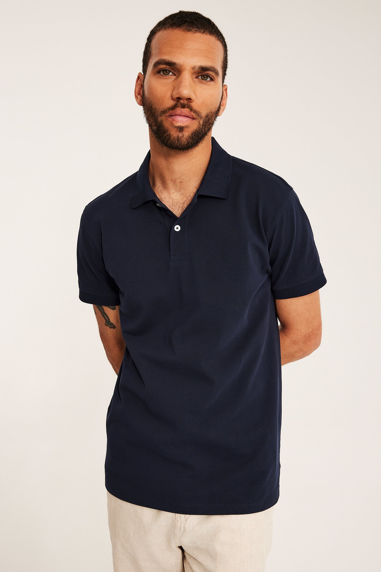 En mann har på seg en mørkeblå bomulls-pikétrøye med to hvite knapper og lysebeige bukser.