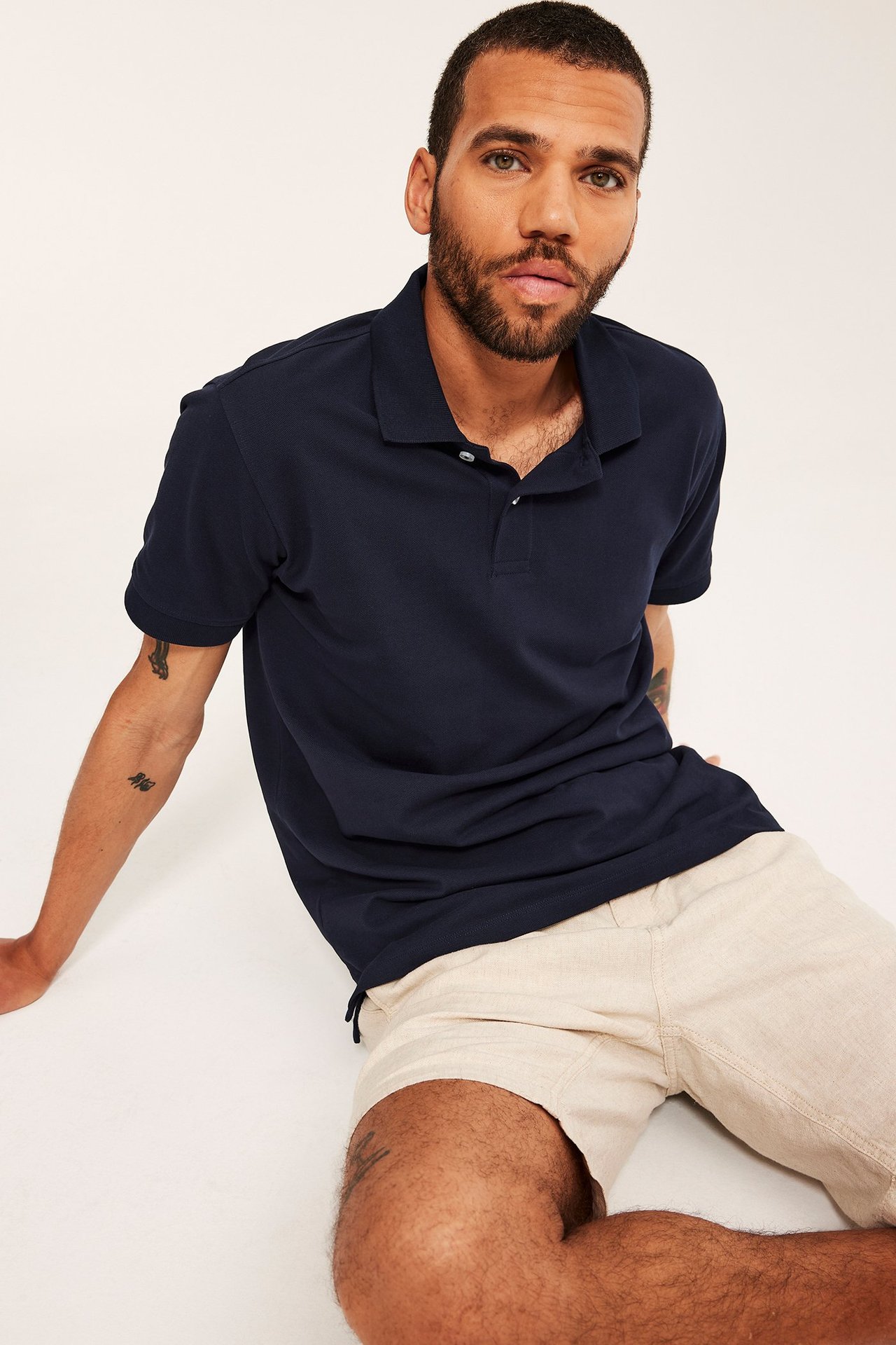 Frontvy: En man i mörkblå pikétröja av bomull med två knappar och ljusbeige shorts.