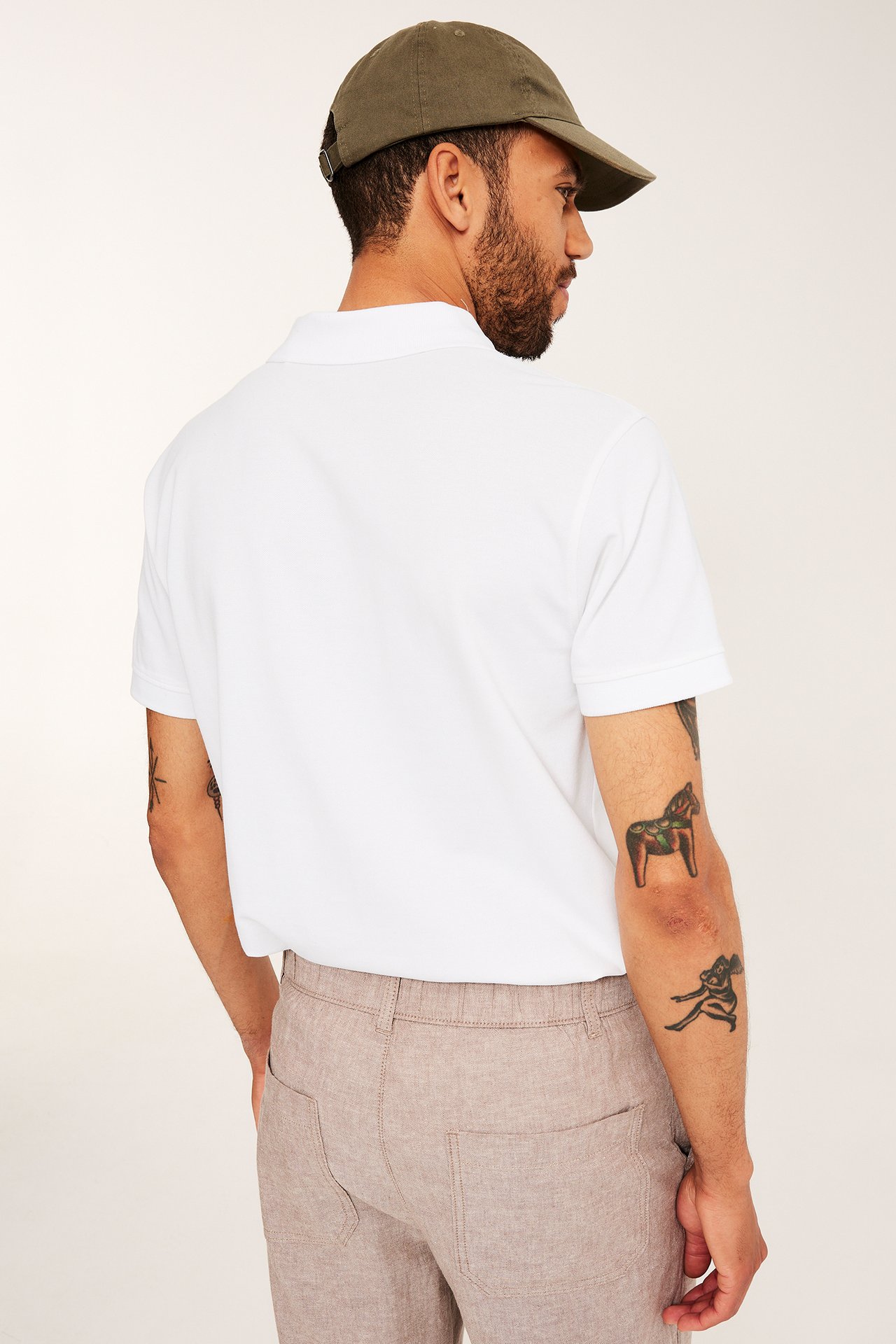 Ryggvy av man i vit kortärmad pikétröja av bomull, en olivgrön keps och beigefärgade byxor.