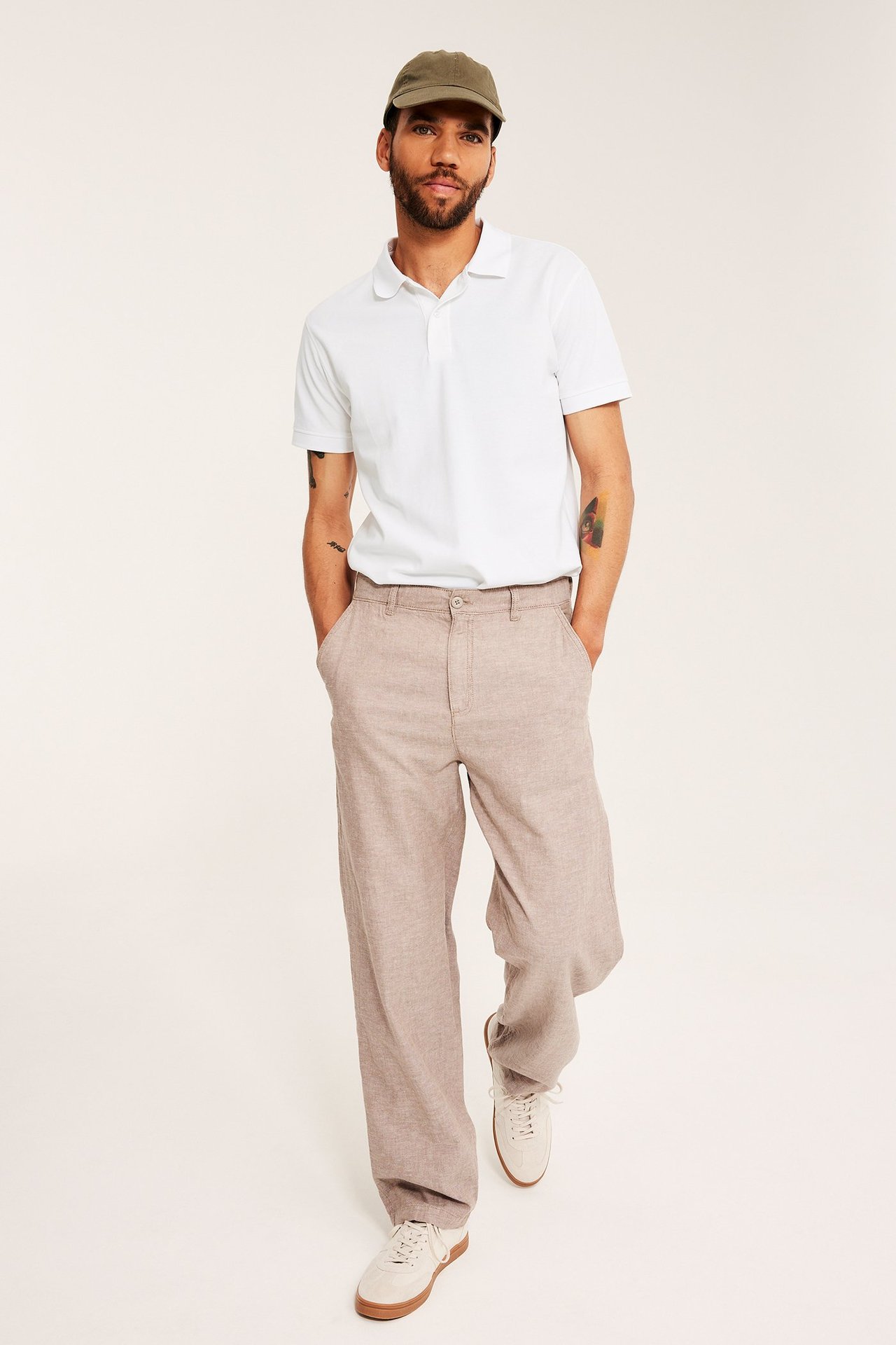 Man i vit bomullspikétröja, beige vida byxor, vita sneakers och olivgrön keps, framifrån.