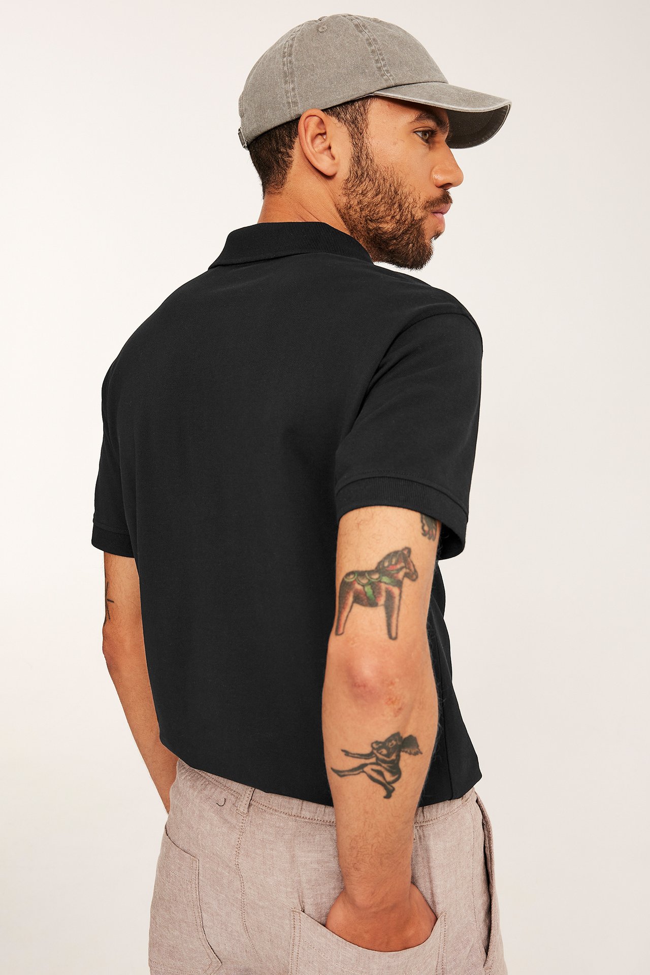 Mies selin-sivulta, yllään musta puuvillainen pikeepaita, harmaa lippalakki ja vaaleanbeiget vapaa-ajan housut.