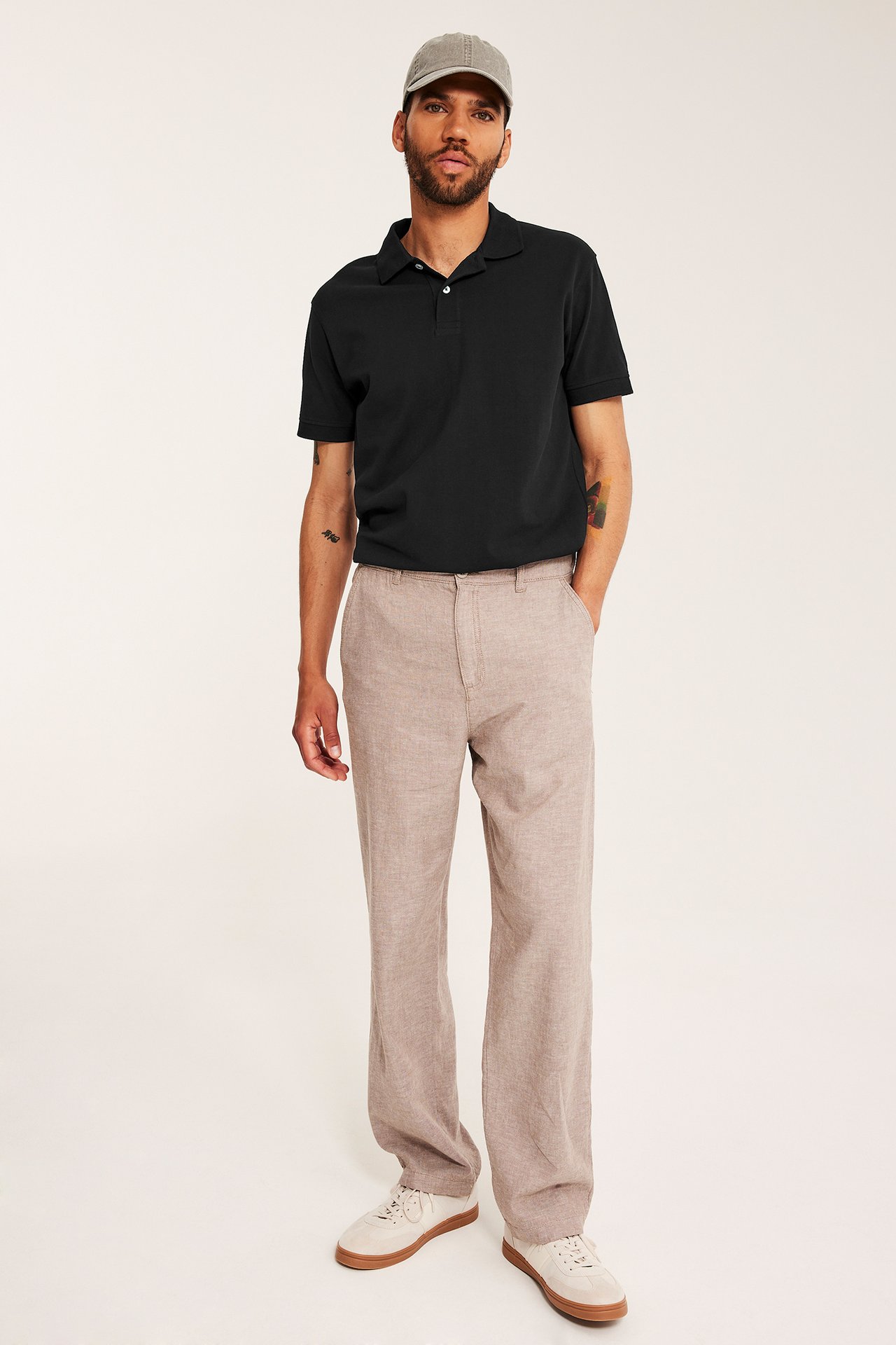 Etunäkymä miehestä: musta pikeepaita, beiget housut, valkoiset tennarit, harmaa lippalakki.