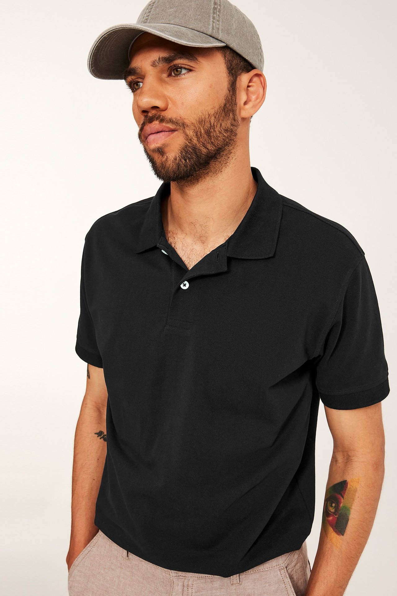 Framsida: man i svart bomullspikétröja med två vita knappar, grå keps och ljusbeiga byxor.