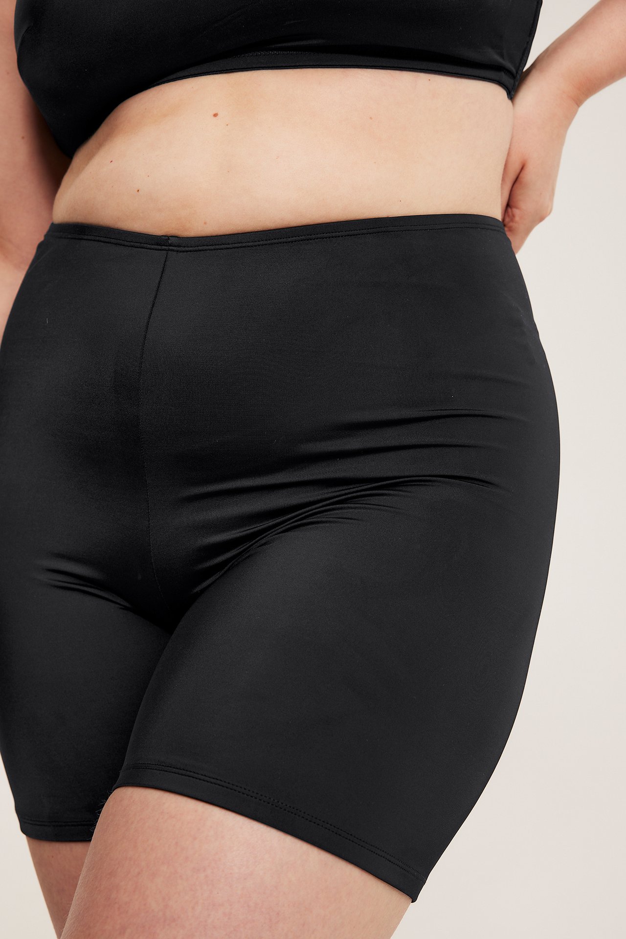 Nærbilde forfra av en kvinne iført svarte, høytlivede badeshorts og en svart bikinitopp.