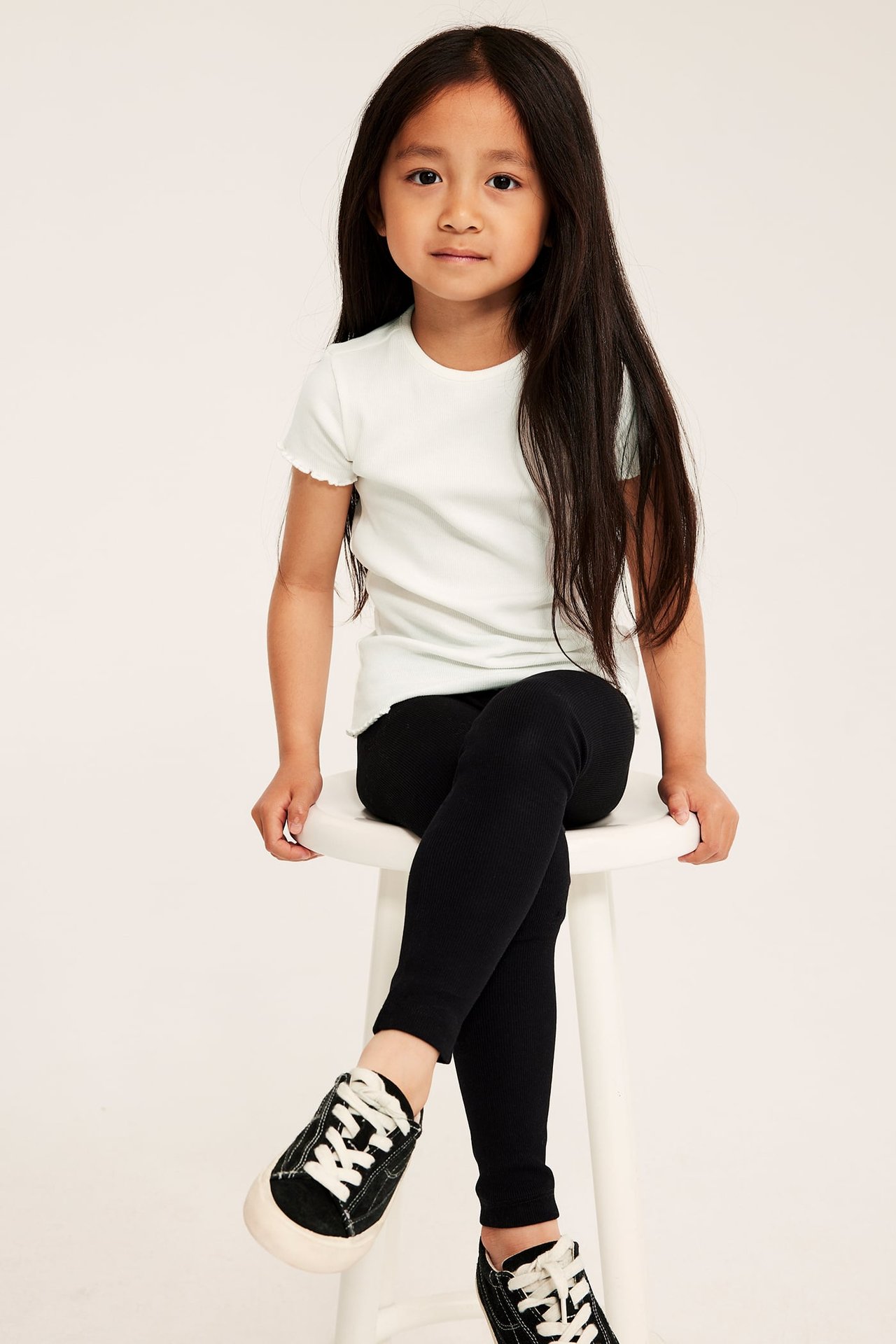 Framifrån: barn i naturvit ribbad kortärmad topp med babylock, svarta ribbade leggings och svarta sneakers.