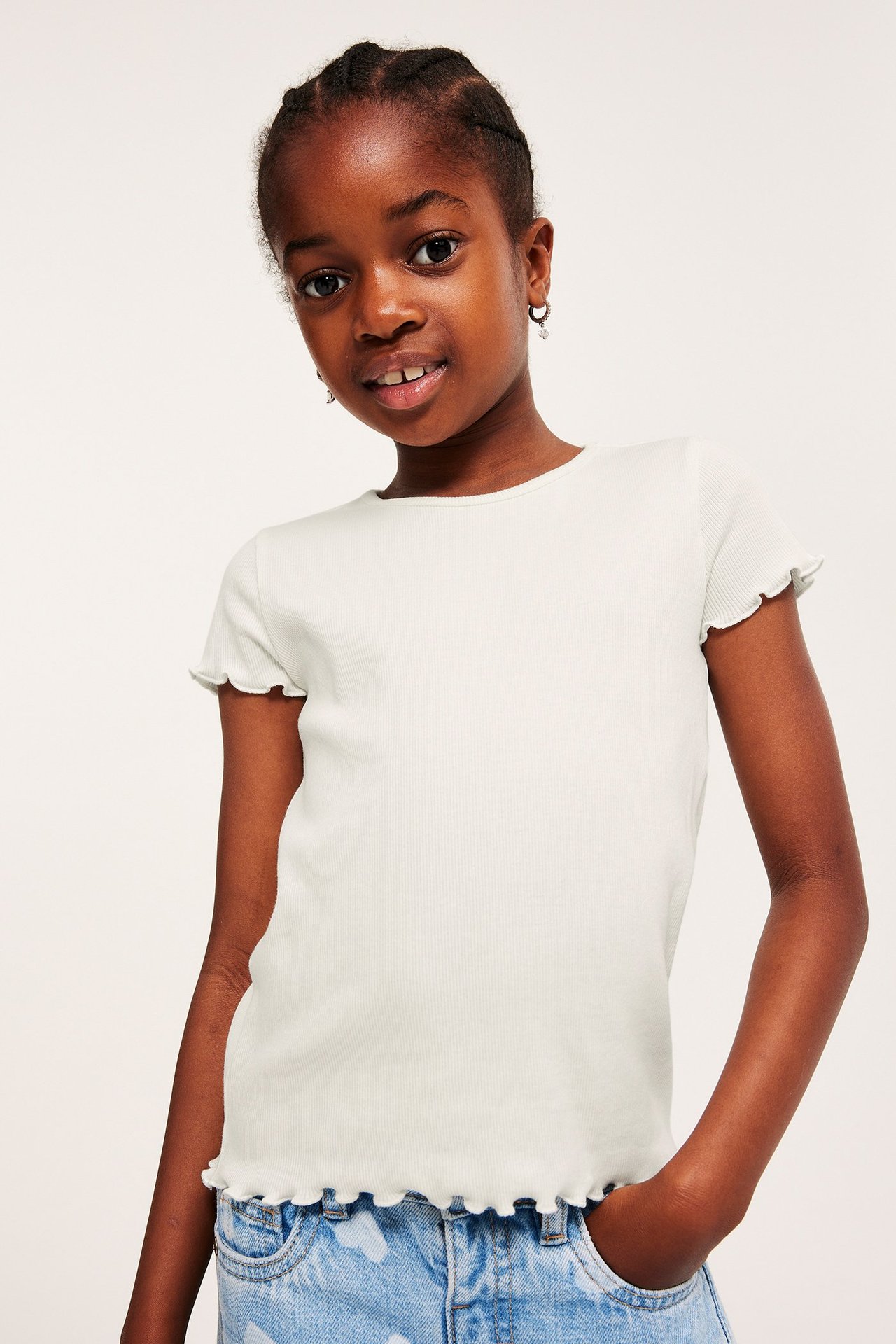 Framifrån, ett barn i en offwhite ribbad kortärmad topp med babylock-kanter och ljusblå jeansshorts.