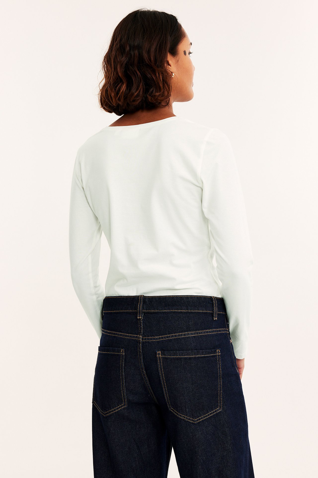 Bakifrån: en kvinna i en offwhite långärmad bastopp med rundhals och mörkblå jeans.