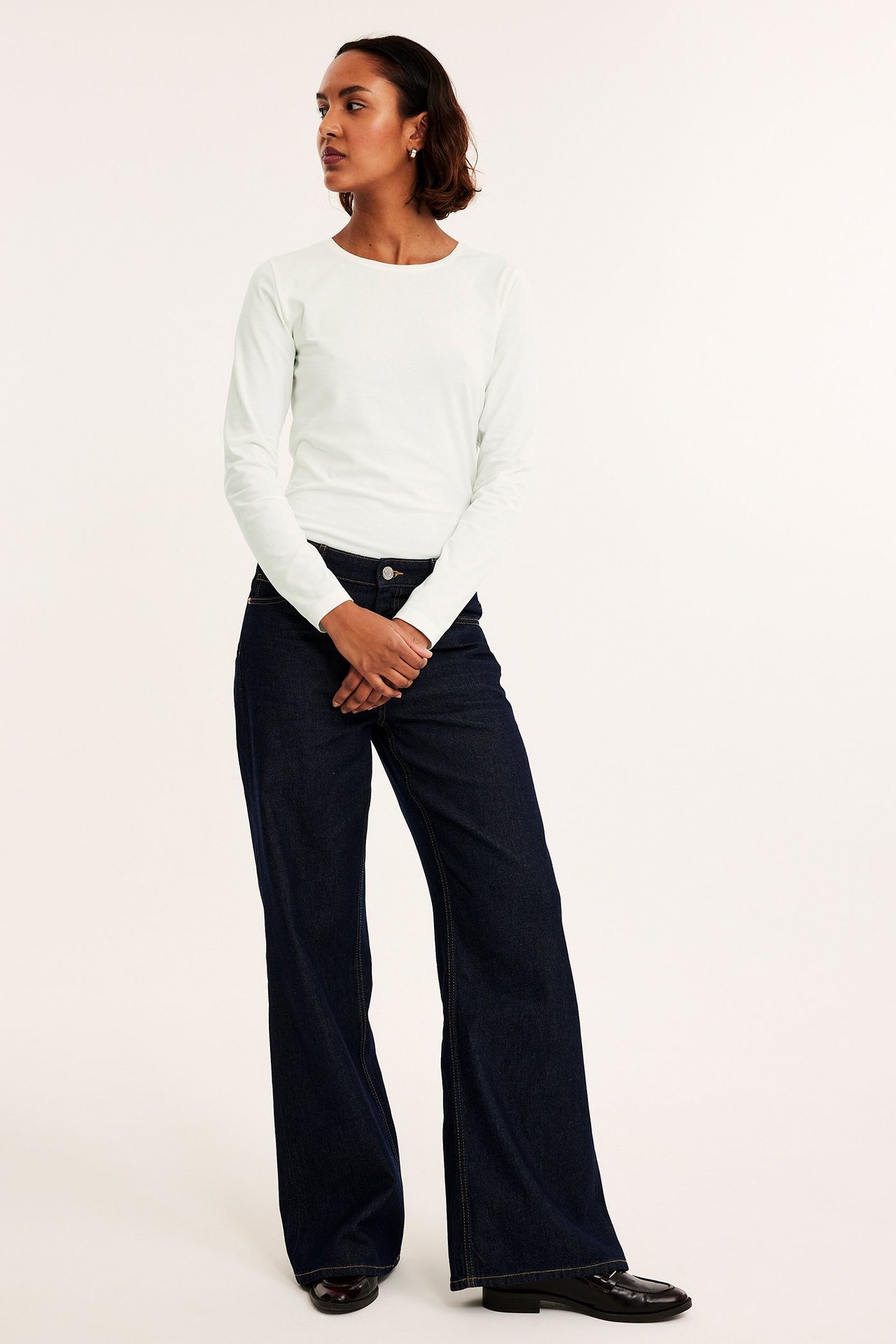Frontbilde: kvinne i offwhite ettersittende langermet topp med rund hals, mørkeblå vide jeans og svarte loafers.