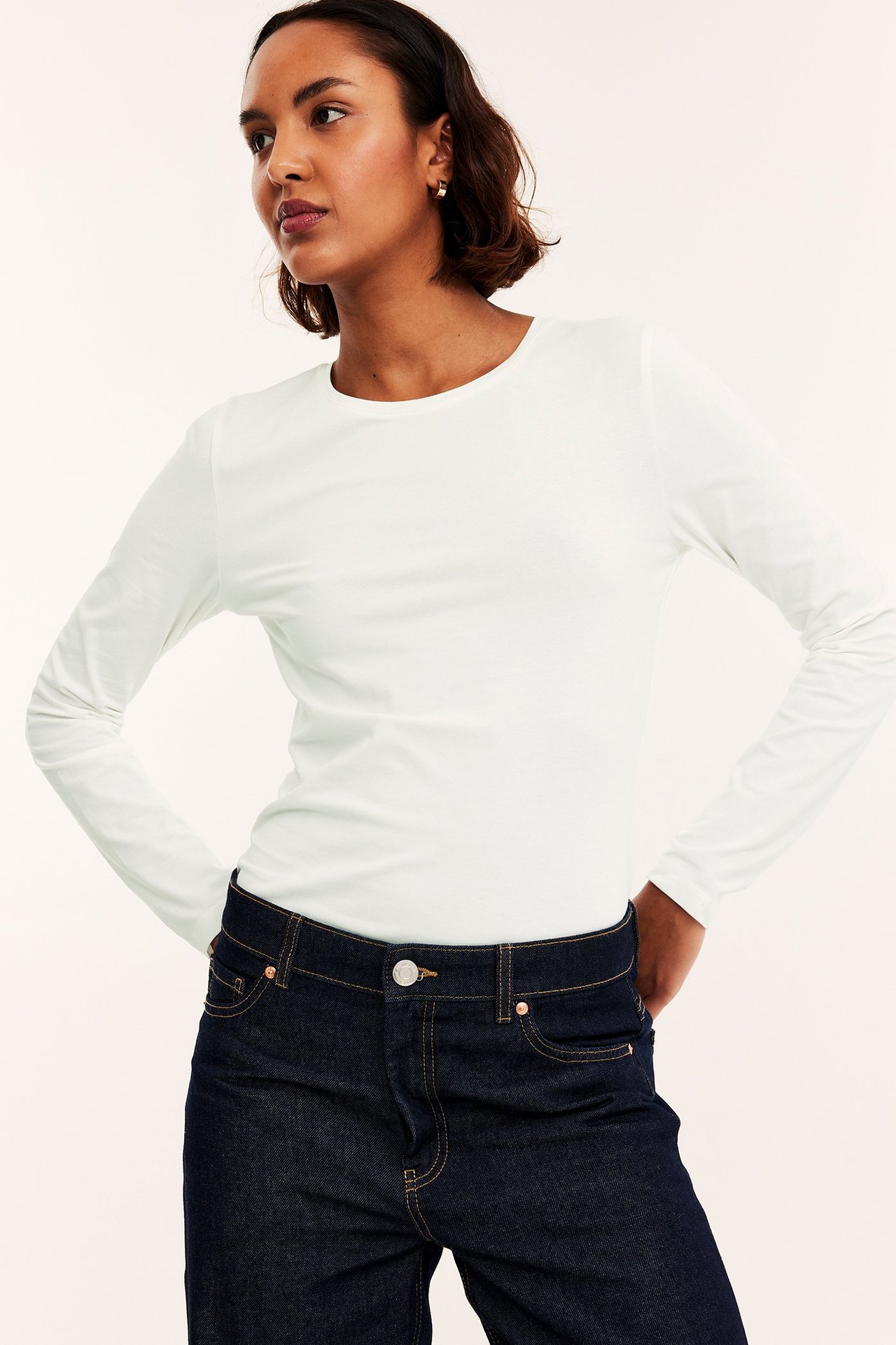Forfra, kvinne i en offwhite langermet basistopp stukket ned i mørkeblå jeans.