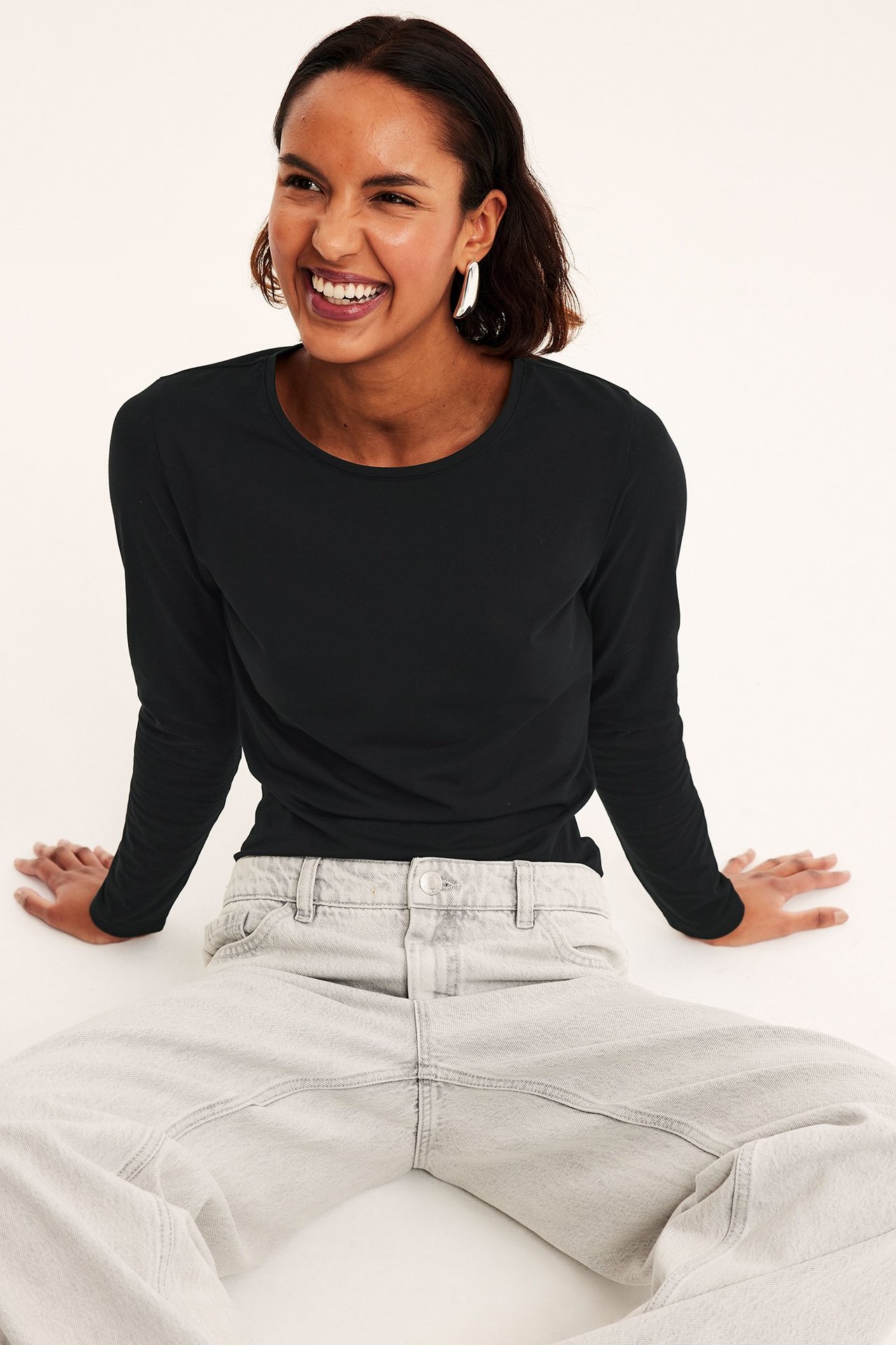 Forfra, kvinne i svart langermet topp, lysegrå jeans og sølvfargede øreringer.