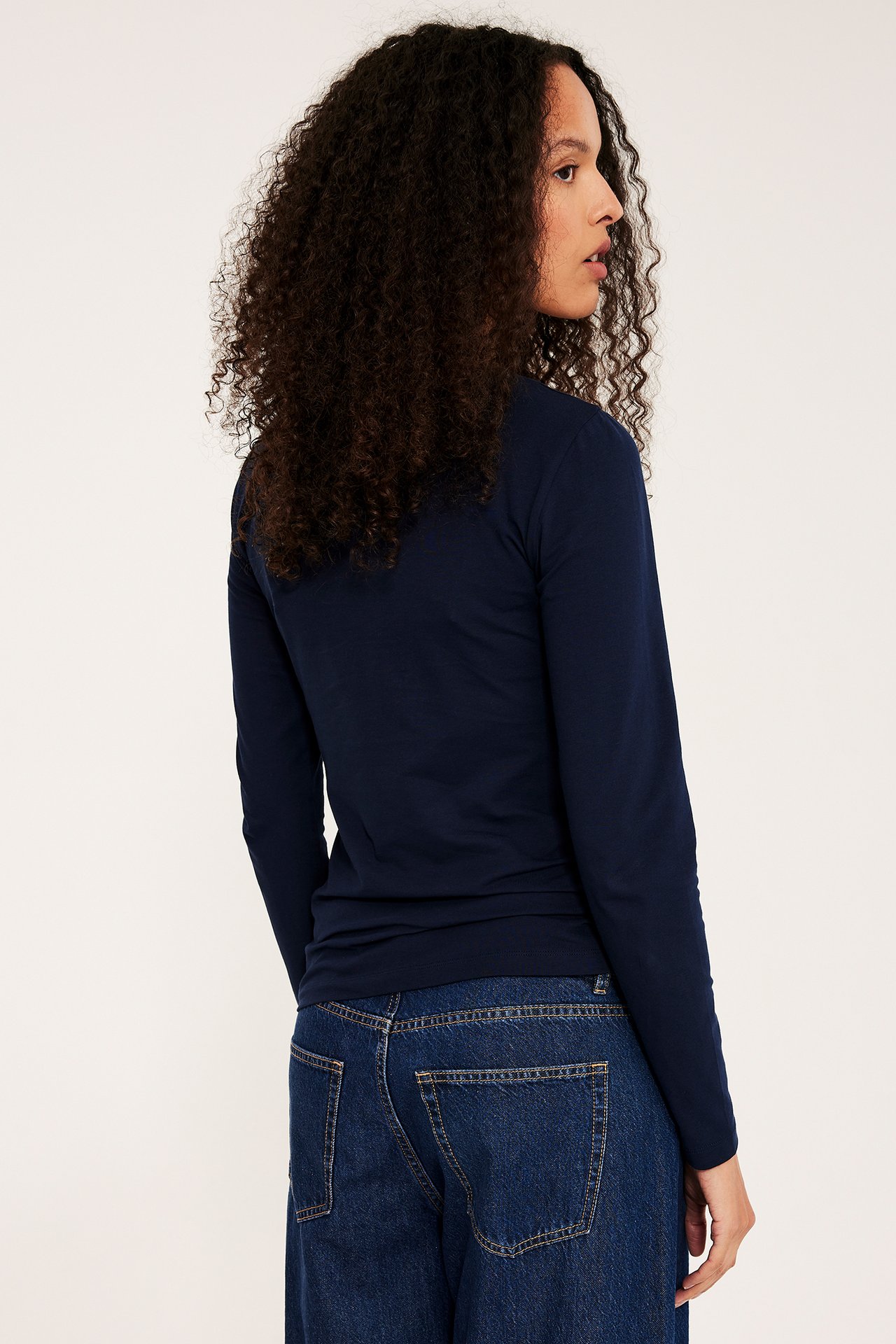Sidovy av kvinna i mörkblå enfärgad långärmad bastopp med rundhals och mörkblå jeans.