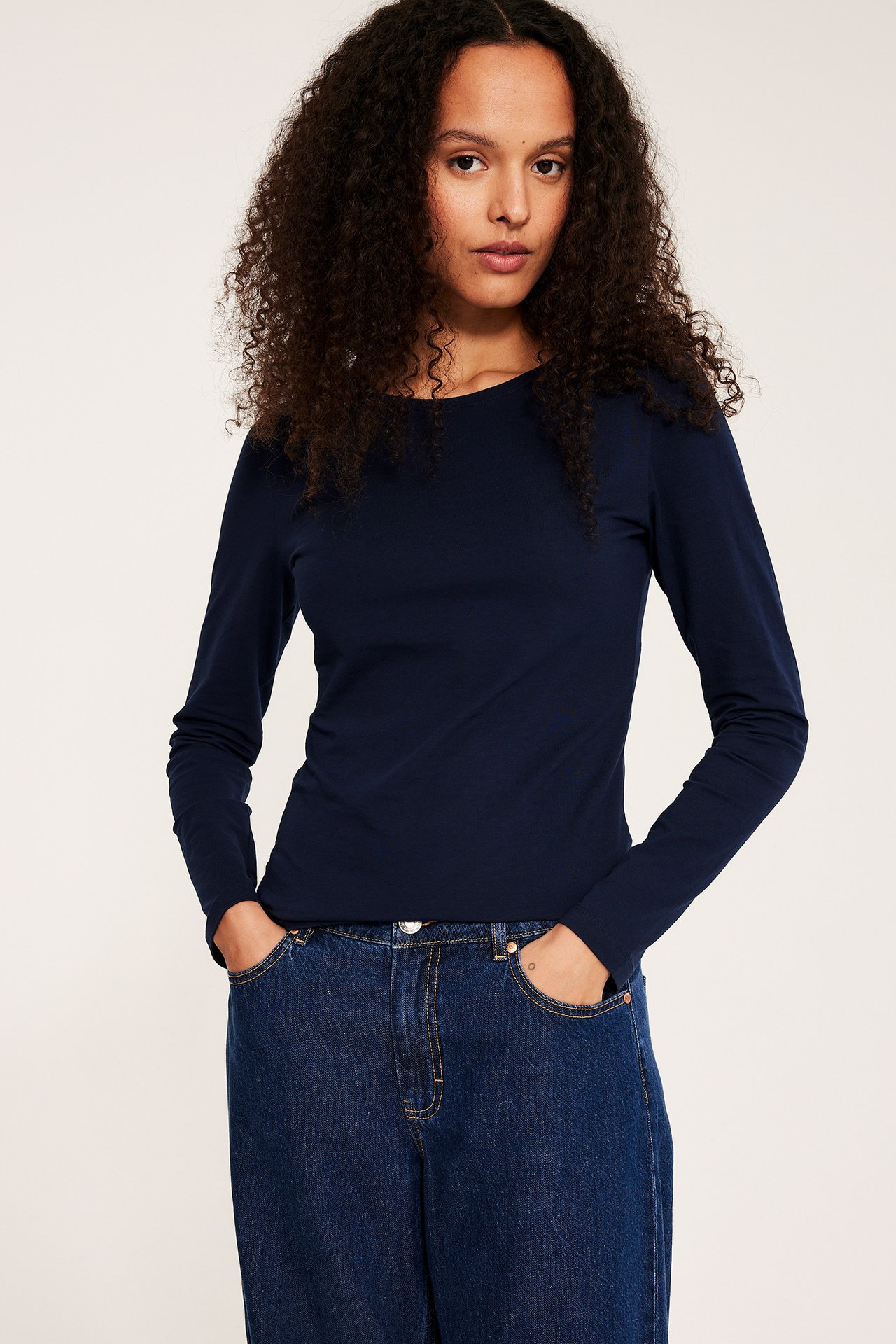 Edestä, nainen yllään tummansininen pitkähihainen peruspusero ja tummansiniset leveälahkeiset farkut.