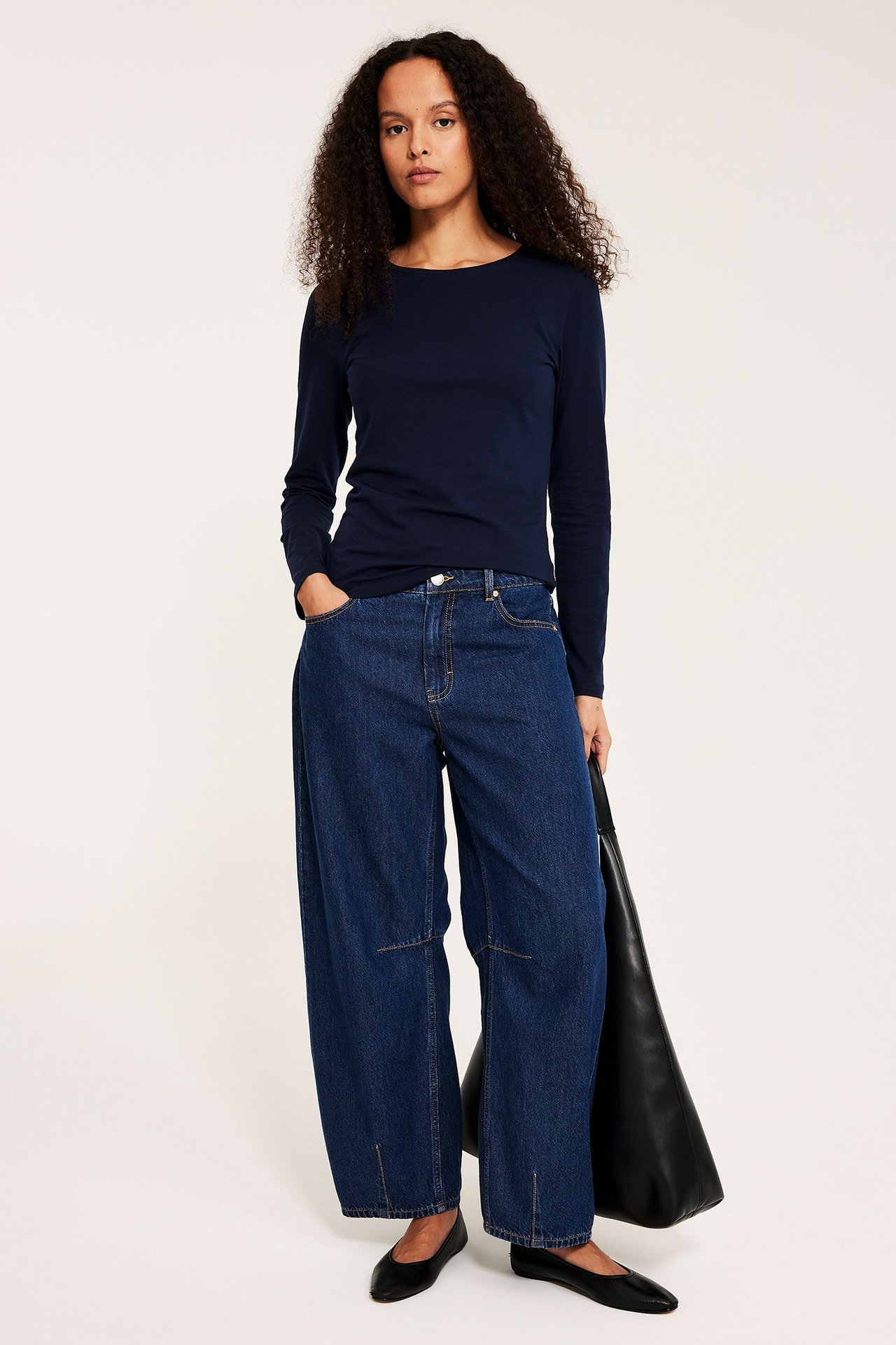 Forfra: kvinne i mørkeblå langermet basistopp, vide jeans, svarte flate sko, holder en svart veske.