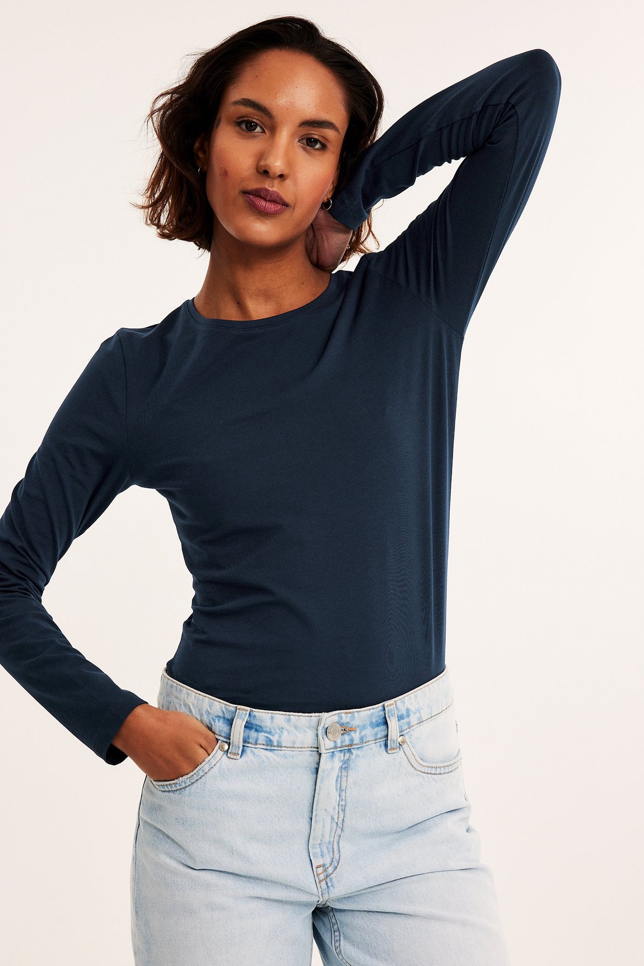 Frontvisning av en kvinne i en mørkeblå langermet rundhalset basistopp og lyseblå jeans.