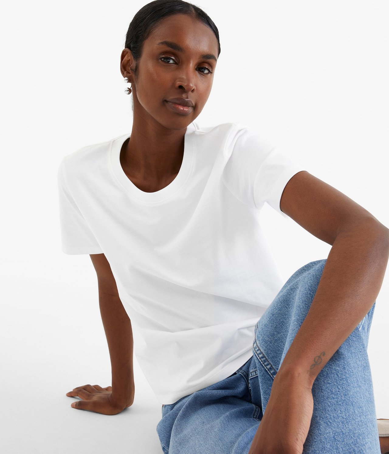 Kvinne i hvit bomulls-T-skjorte med rett passform, blå jeans og sølvfargede øredobber, forfra.
