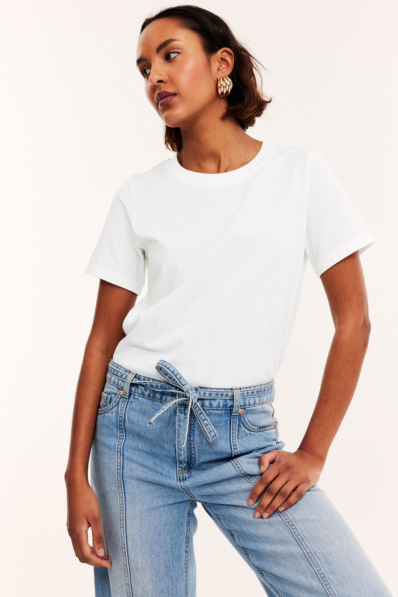 Framifrån, kvinna i offwhite bomulls-t-shirt, ljusblå jeans med knytmidja och guldfärgade örhängen.