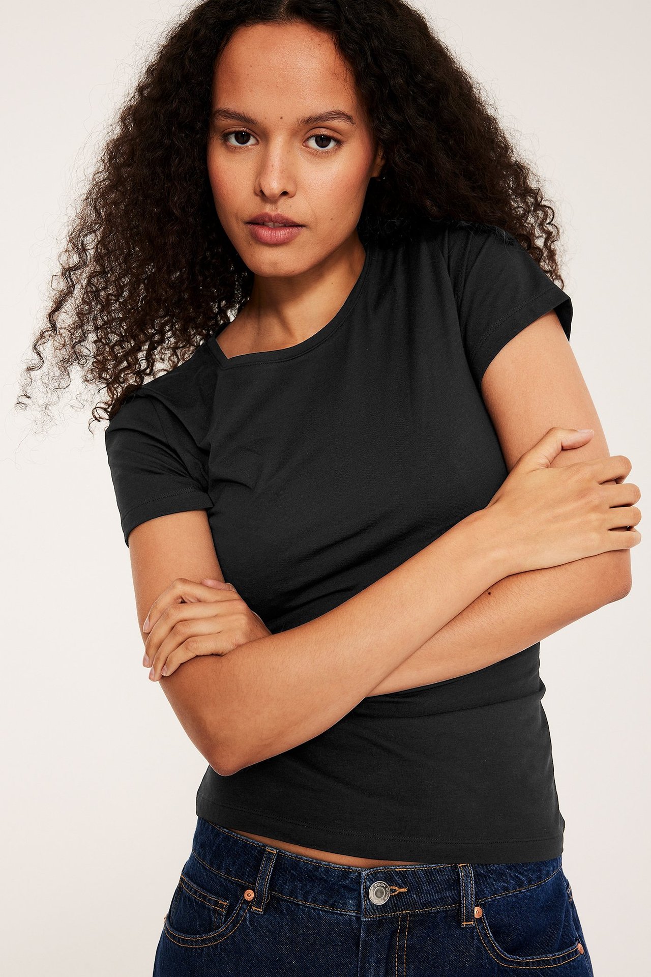 Frontbilde av en kvinne i en svart kortermet basistopp med rund hals og mørkeblå jeans.
