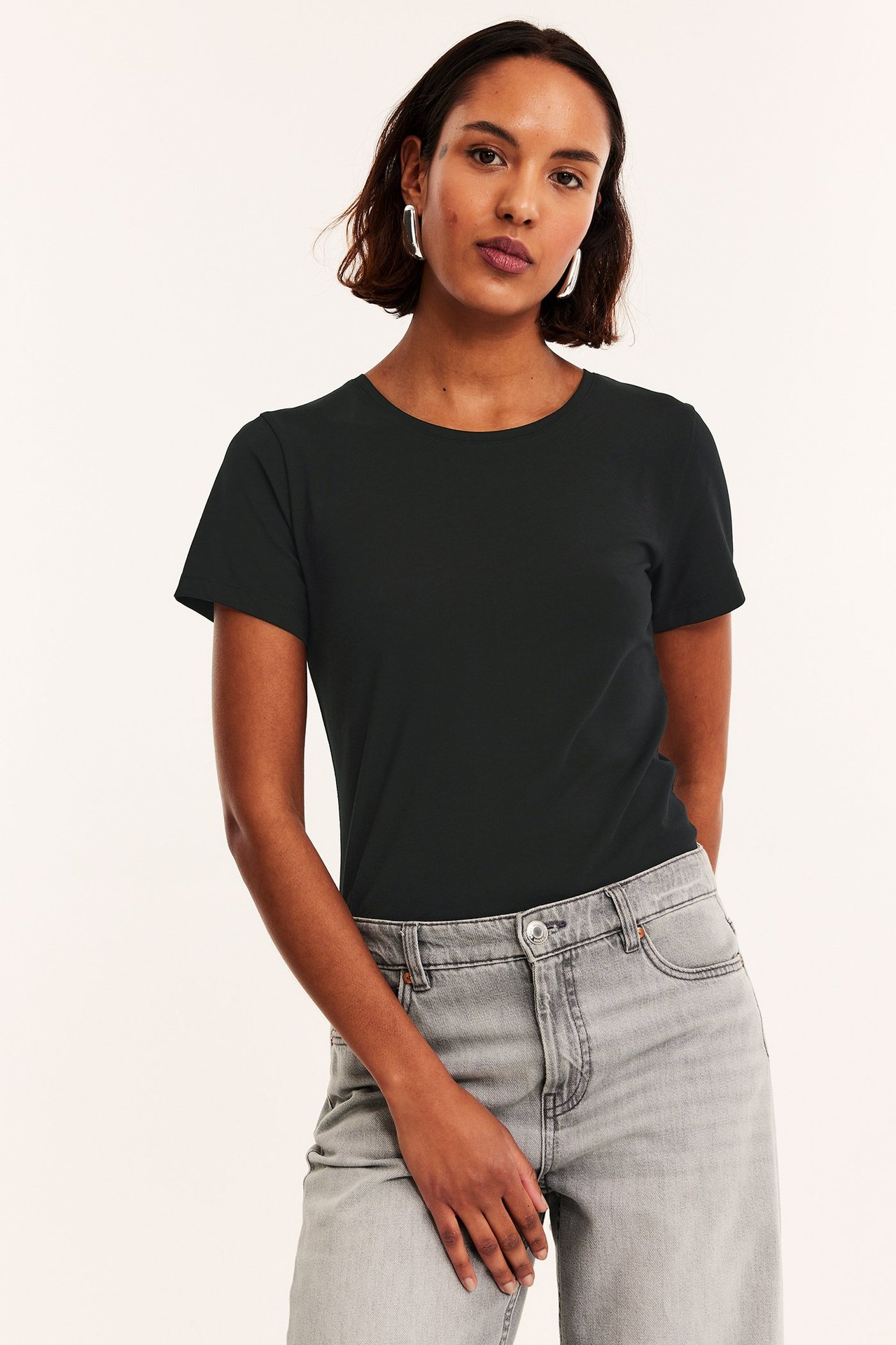 Frontbilde av kvinne i svart kortermet rundhalset basistopp, grå jeans og sølvfargede øredobber.