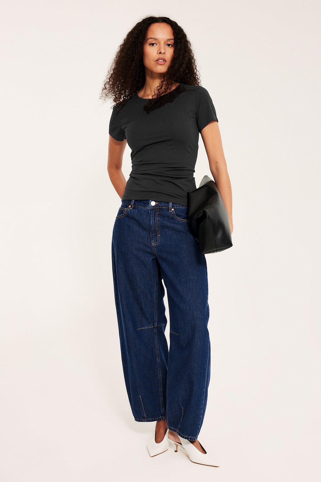 Frontvisning: Kvinne i ettersittende svart kortermet topp, vide mørkeblå jeans, hvite hæler, holder svart veske.