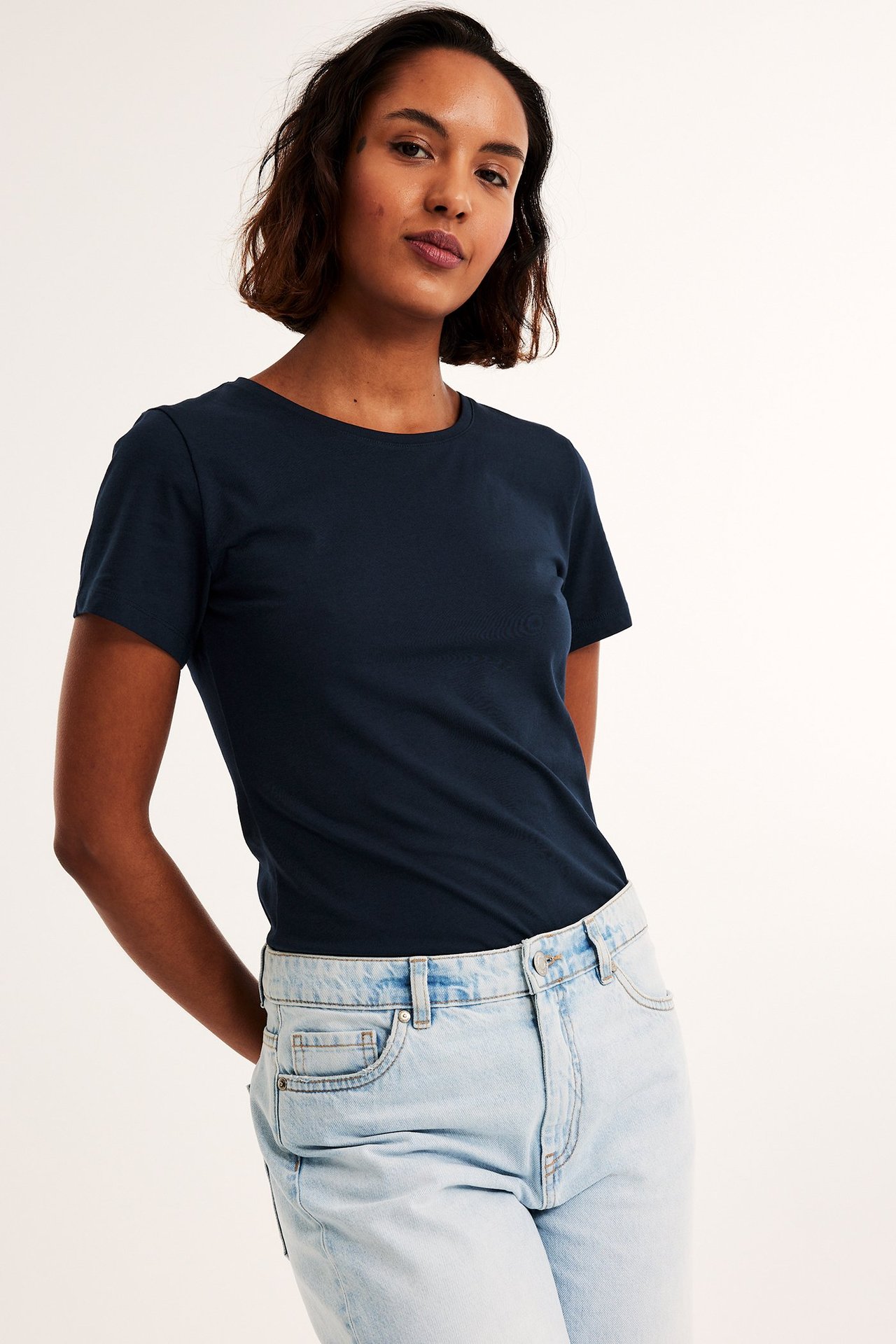 Forfra: kvinne i mørkeblå kortermet basistopp med rund hals og lyseblå jeans.